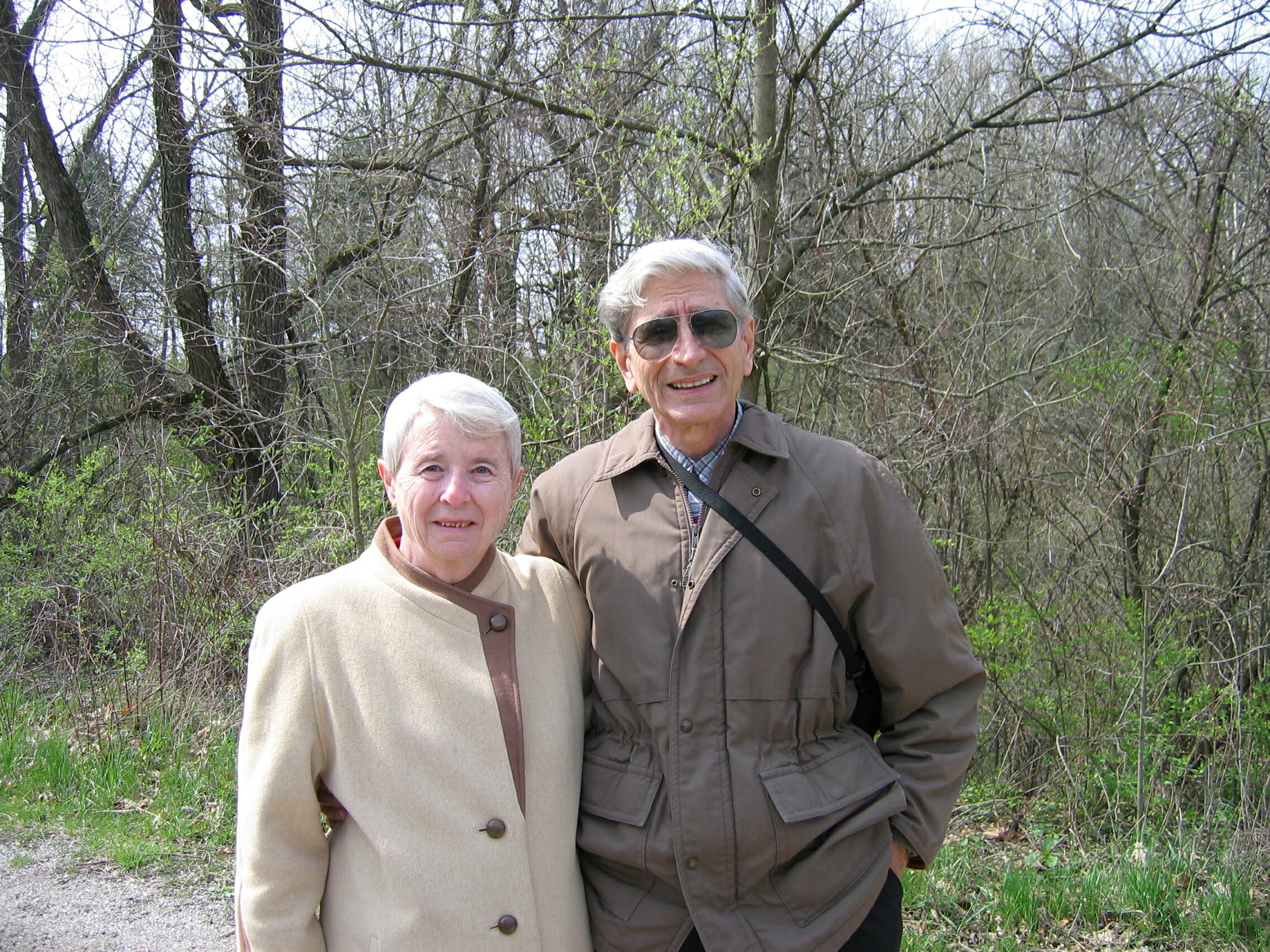 Two seniors standing outdoors in a wooded area, woman in a beige coat and man in a brown jacket posing together.