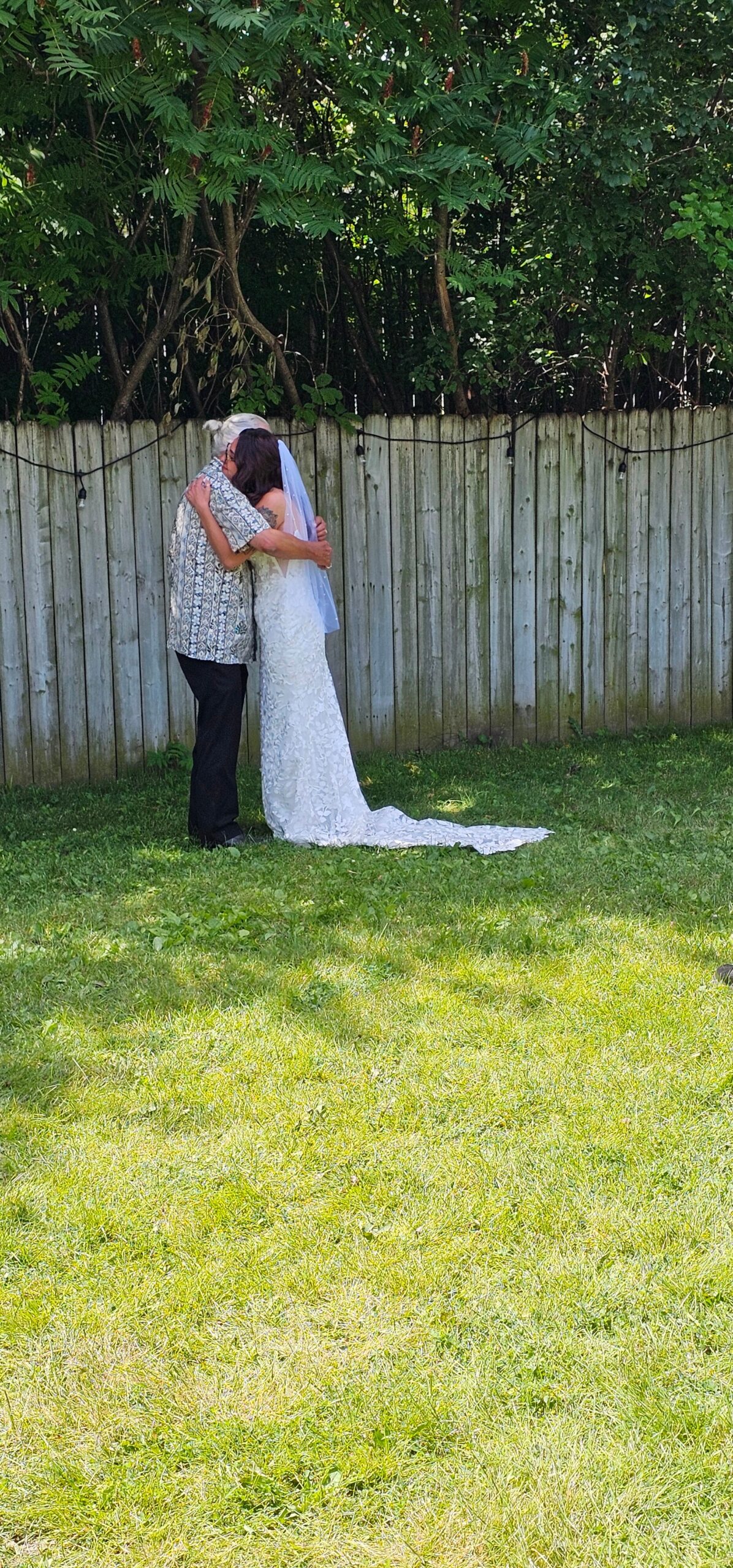 Bride in a lace wedding dress hugs an older man in a patterned shirt in a backyard, near a wooden fence with string lights and green trees behind.