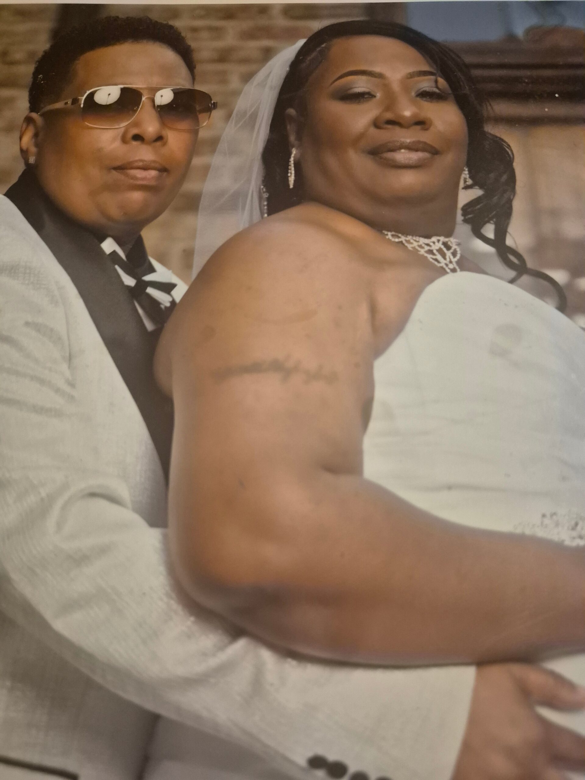 Bride in a white wedding gown with veil and jewelry, smiling slightly, standing beside a man in a light suit and sunglasses.