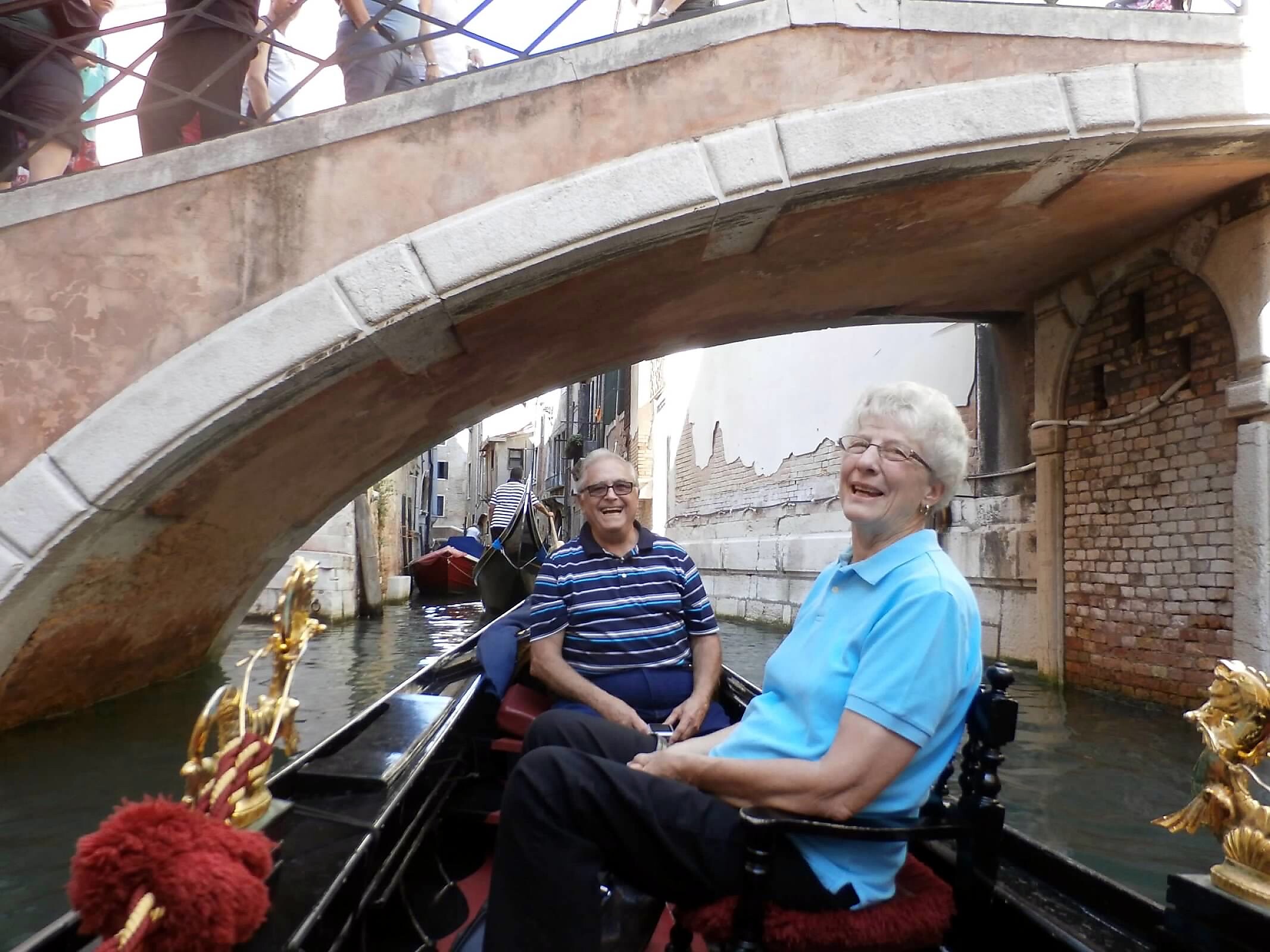 Two elderly people smile while sitting in a narrow canal boat under a brick arch bridge, with decorative red tassels and gold ornaments on the boat.