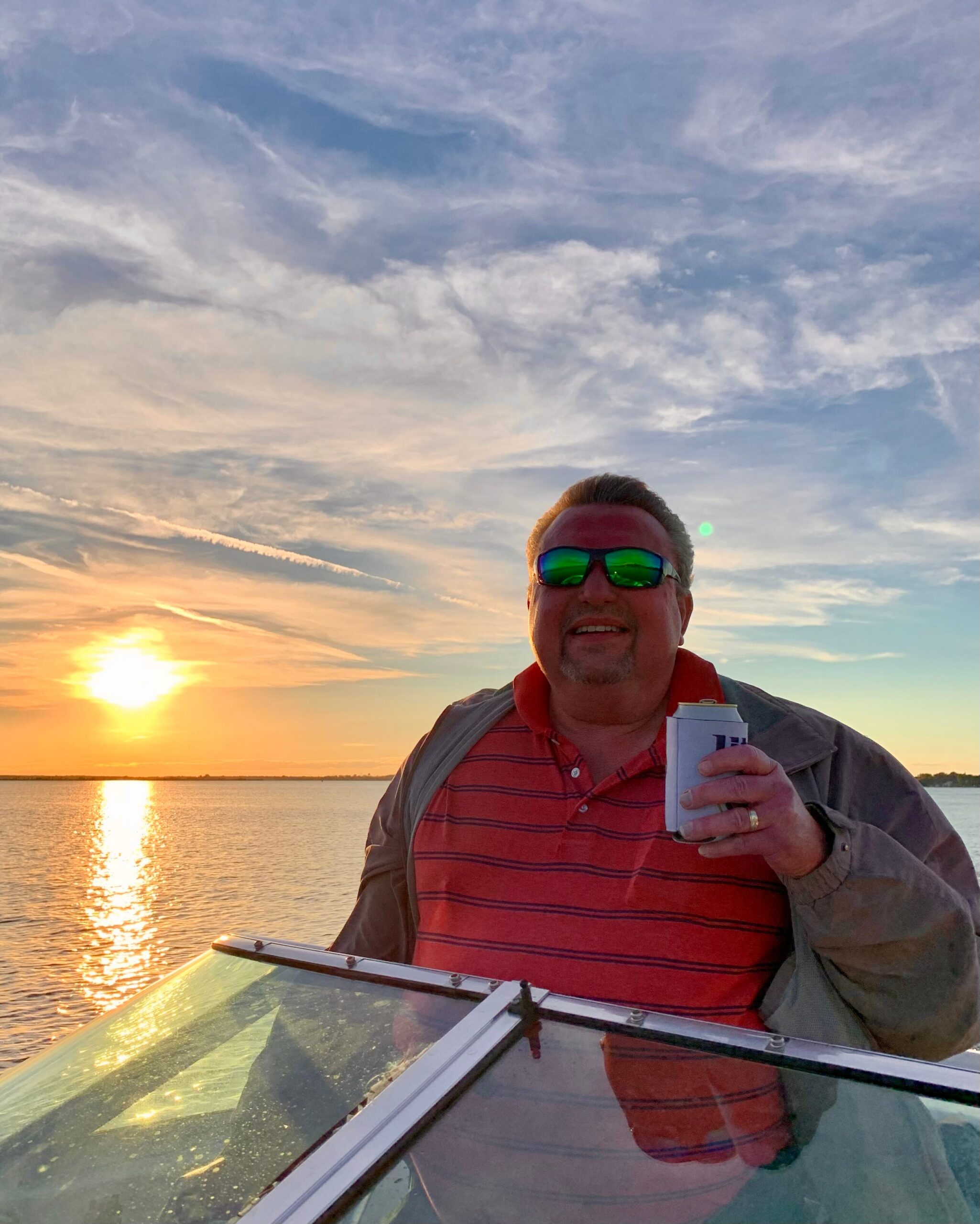 Man on a boat at sunset, wearing a red polo shirt and sunglasses, holding a white booklet or notepad against a reflective water scene.