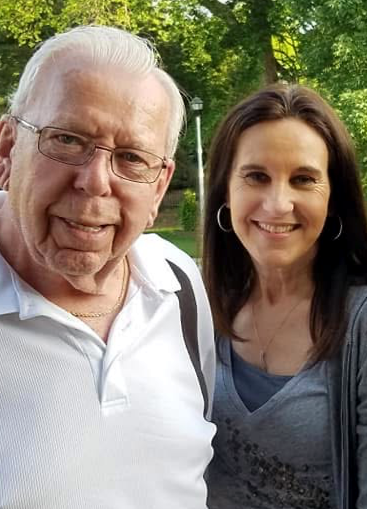 Two adults posing outdoors and smiling, with greenery in the background.
