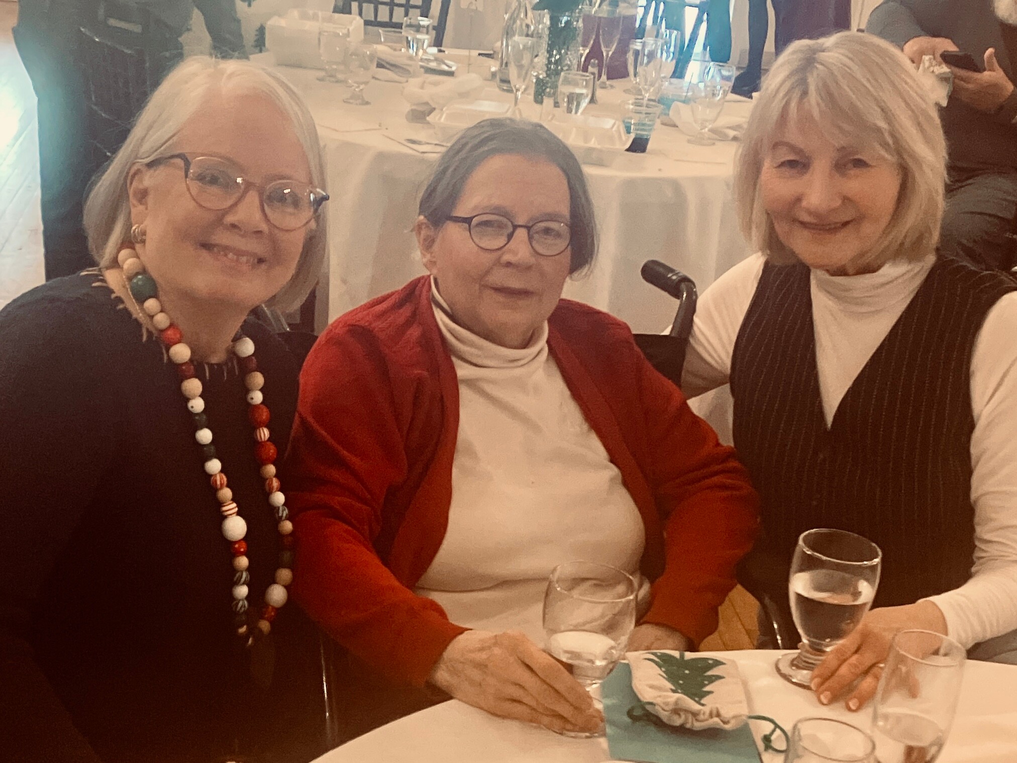 Three smiling older women sit around a dining table with glasses and a wrapped gift bag in the foreground. Left wears a beaded necklace, middle wears glasses and a red cardigan, right wears a striped vest.
