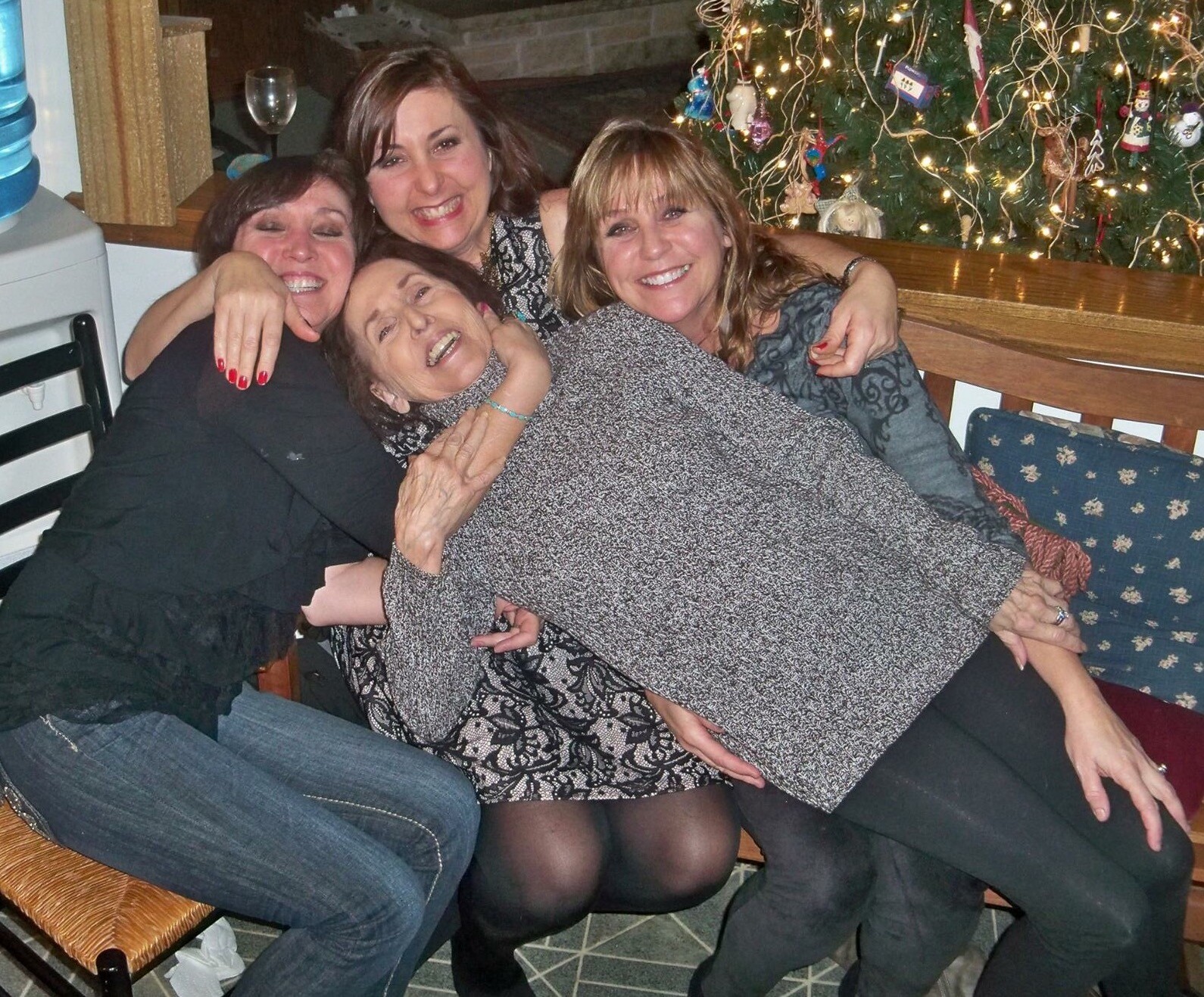 Four women hugging and smiling on a bench with a Christmas tree in the background.