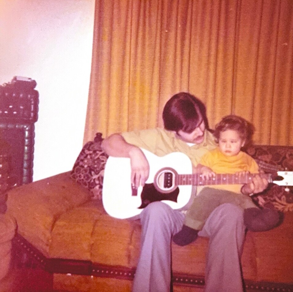 Adult and child sit on a patterned couch while the adult guides the child in playing a white acoustic guitar in a retro living room. **(informative)**