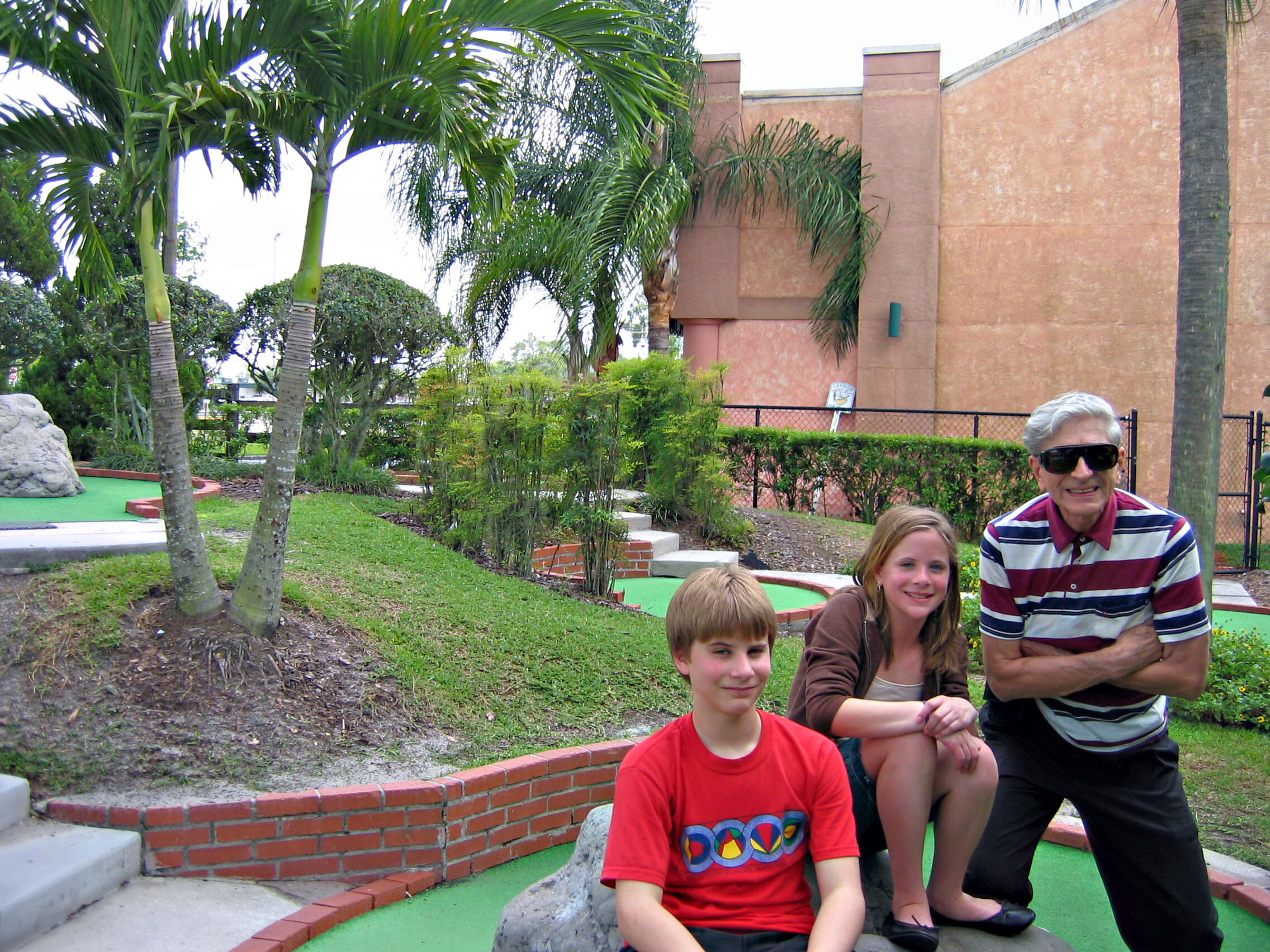 A group of smiling people posing outdoors on a mini-golf course with palm trees and a brick wall in the background.