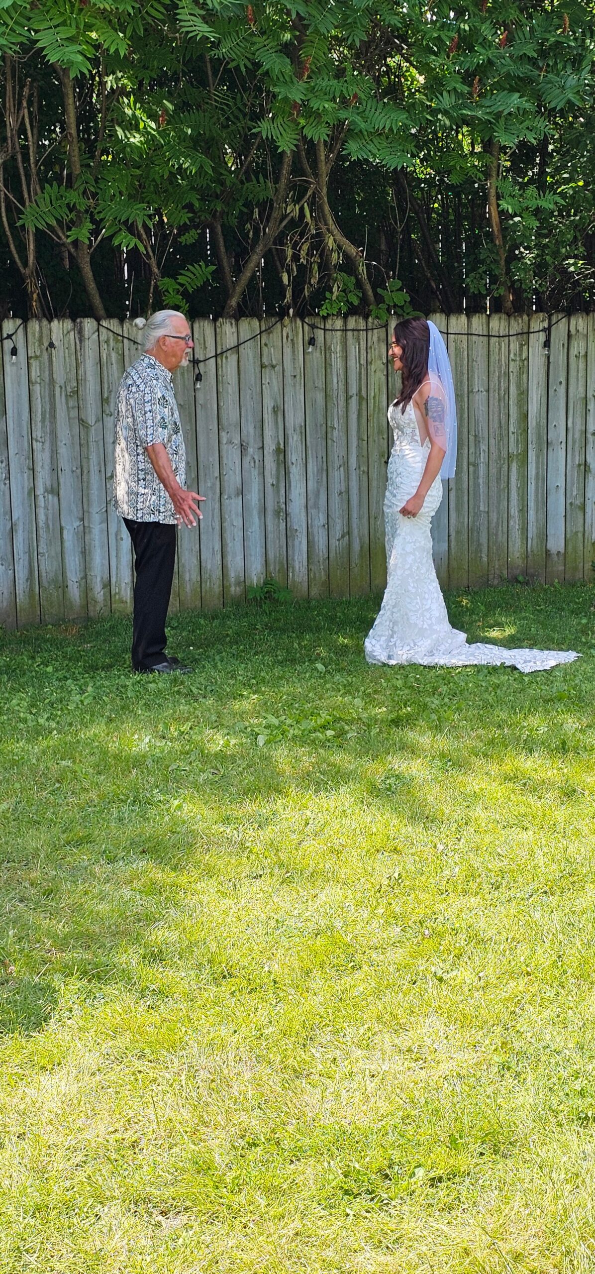 Older man in a patterned short-sleeve shirt talks with a bride in a white lace wedding gown and veil in a backyard.