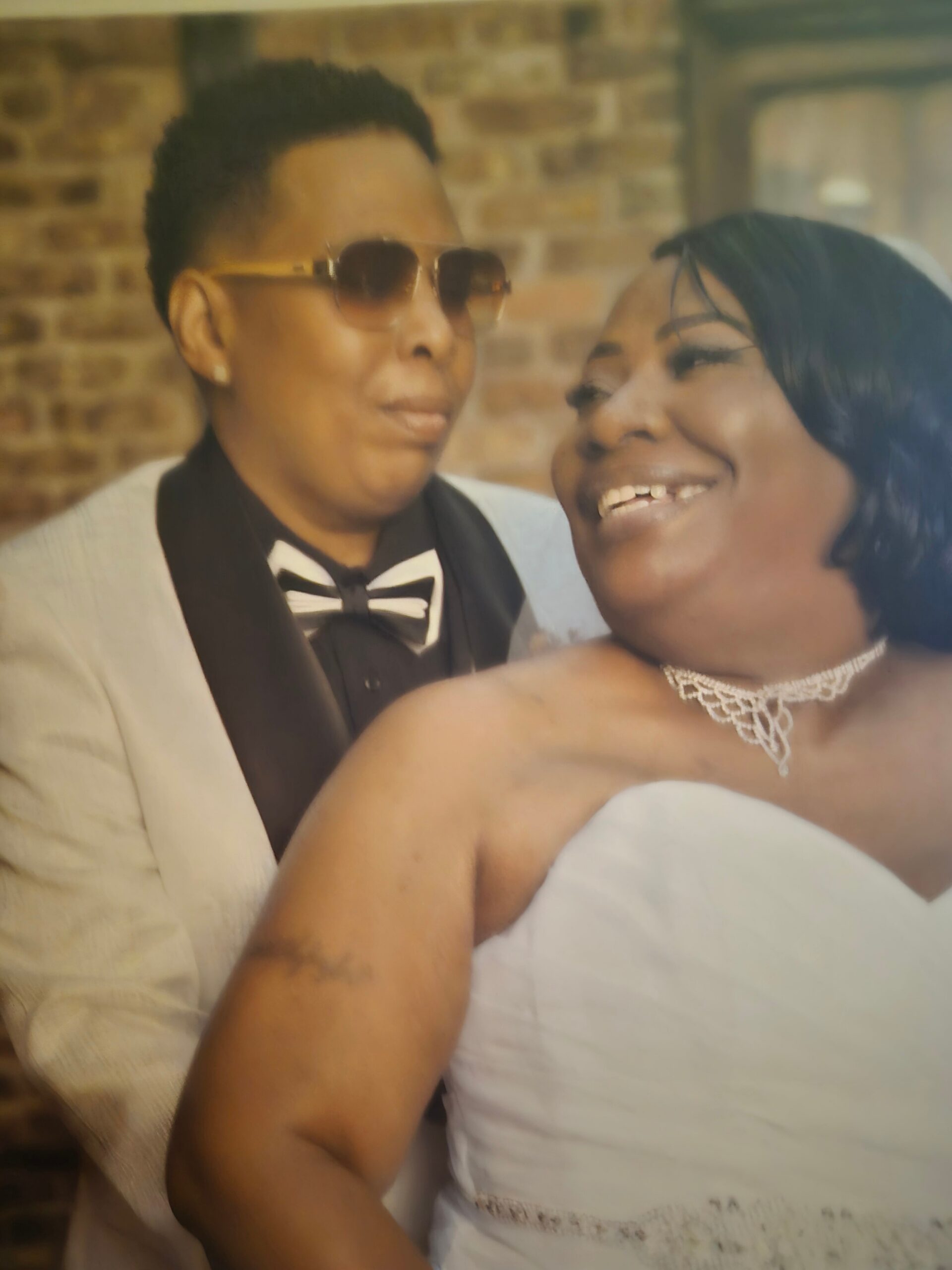 Couple at a wedding: man in a tan suit and bow tie wearing sunglasses, beside a smiling woman in a white dress with a lace necklace against a brick wall backdrop.