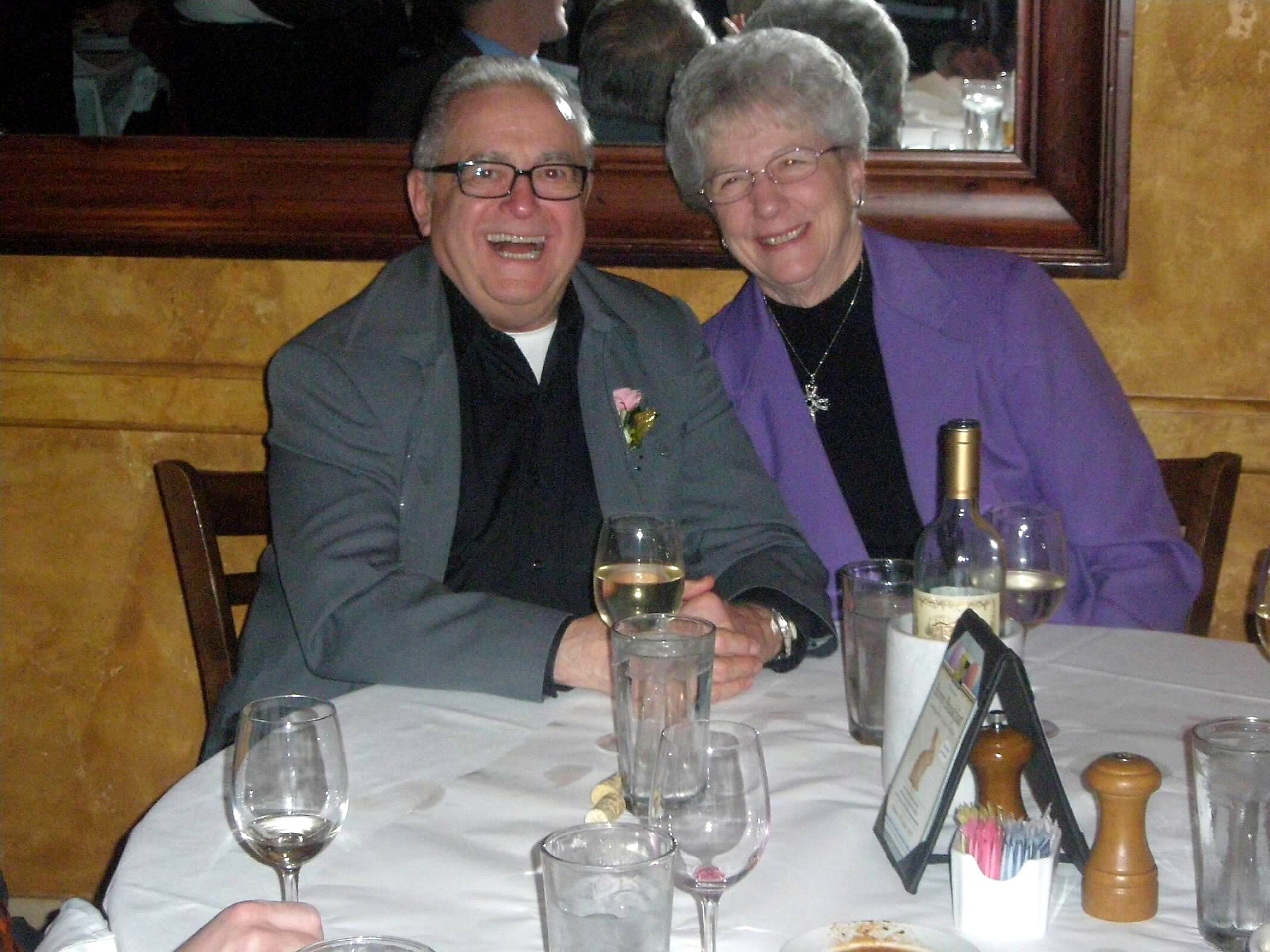 Two smiling older adults sit at a restaurant table, man in gray jacket and woman in purple blazer, with wine glasses nearby.