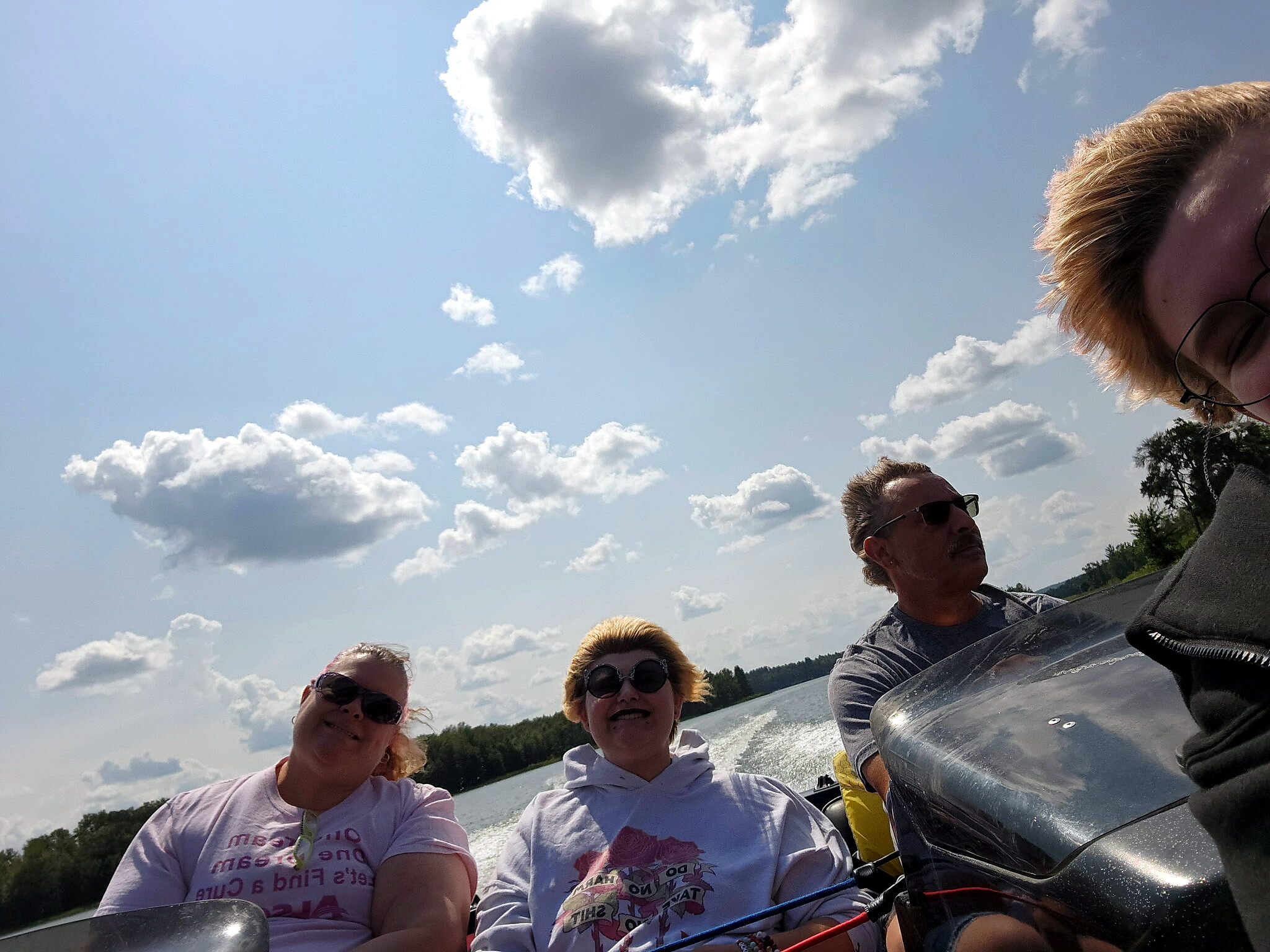 Group of friends on a boat taking a sunny daytime selfie, blue sky with clouds and lake in the background, wearing casual shirts and sunglasses.