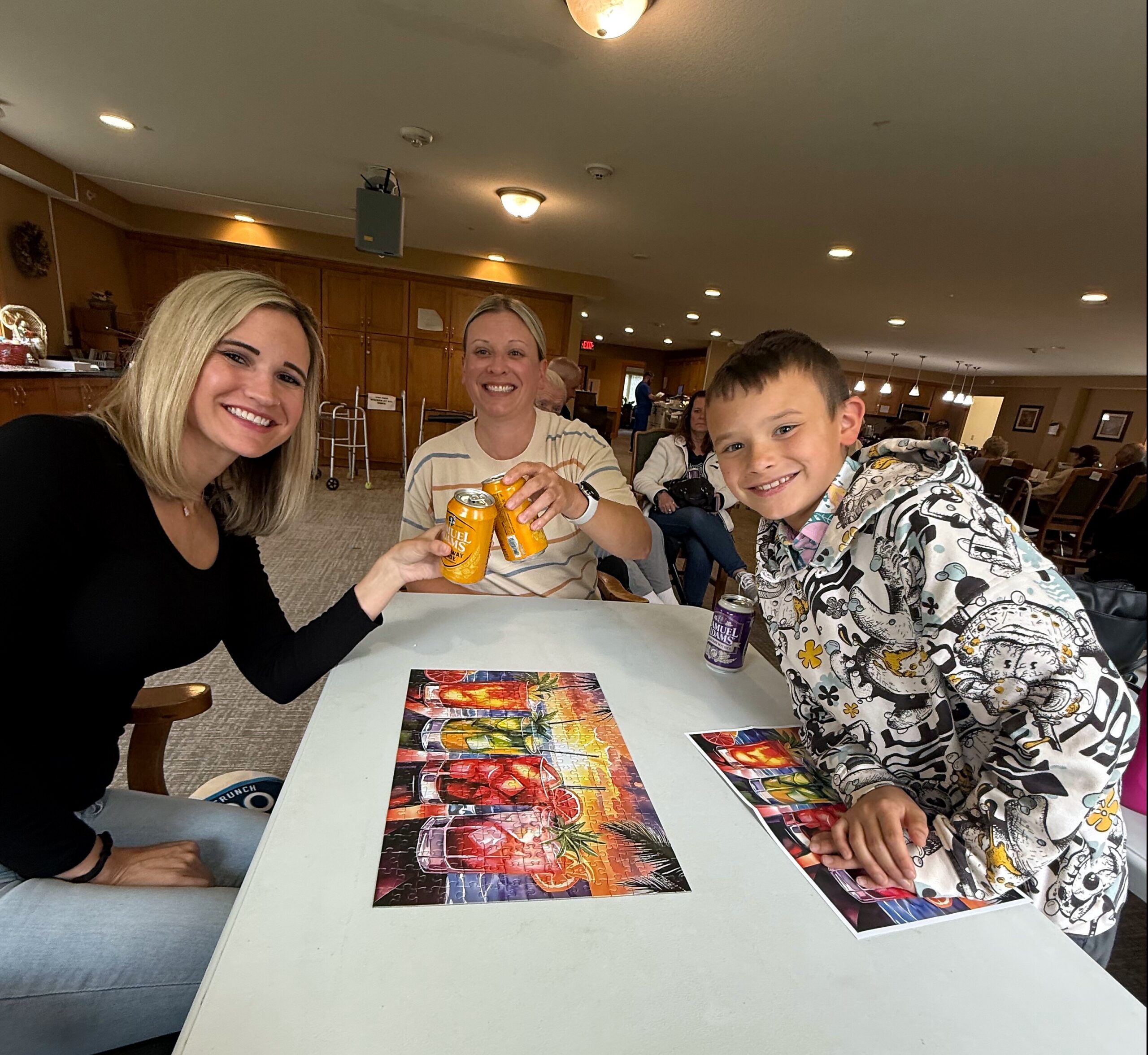 Three smiling people sit around a table with colorful jigsaw puzzle cards spread out and drinks in hand in a bright indoor room.