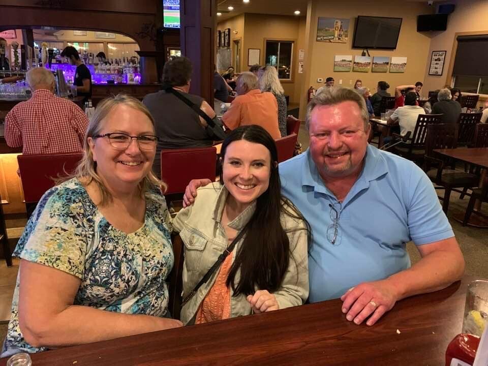 Three adults at a busy restaurant table smiling for the photo: two women (one with patterned blouse, one with denim jacket) and a man in a light blue polo.