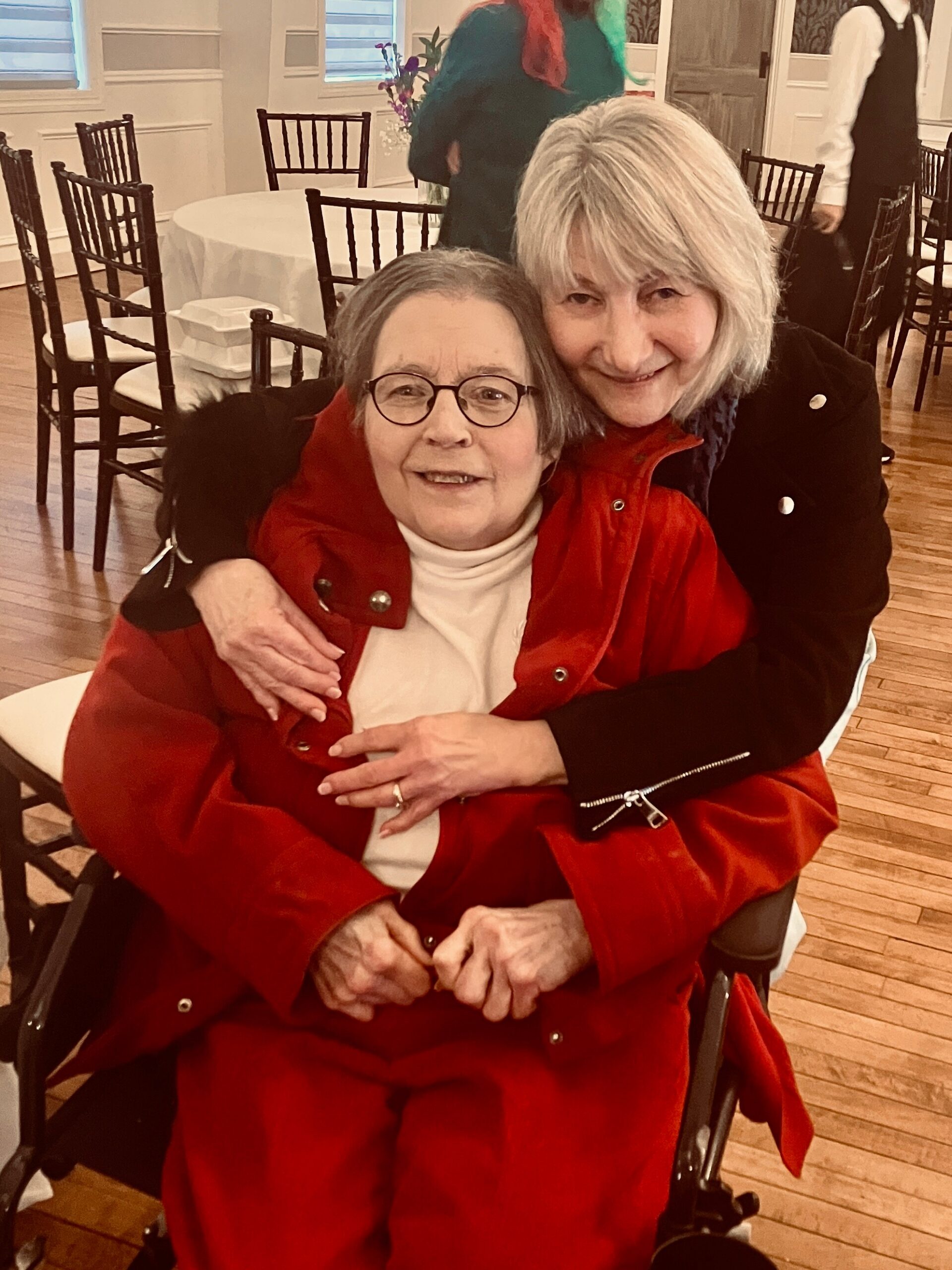 Two smiling elderly women indoors; one seated in a wheelchair wearing a red coat and glasses, the other stands behind her with a hug at a social event.