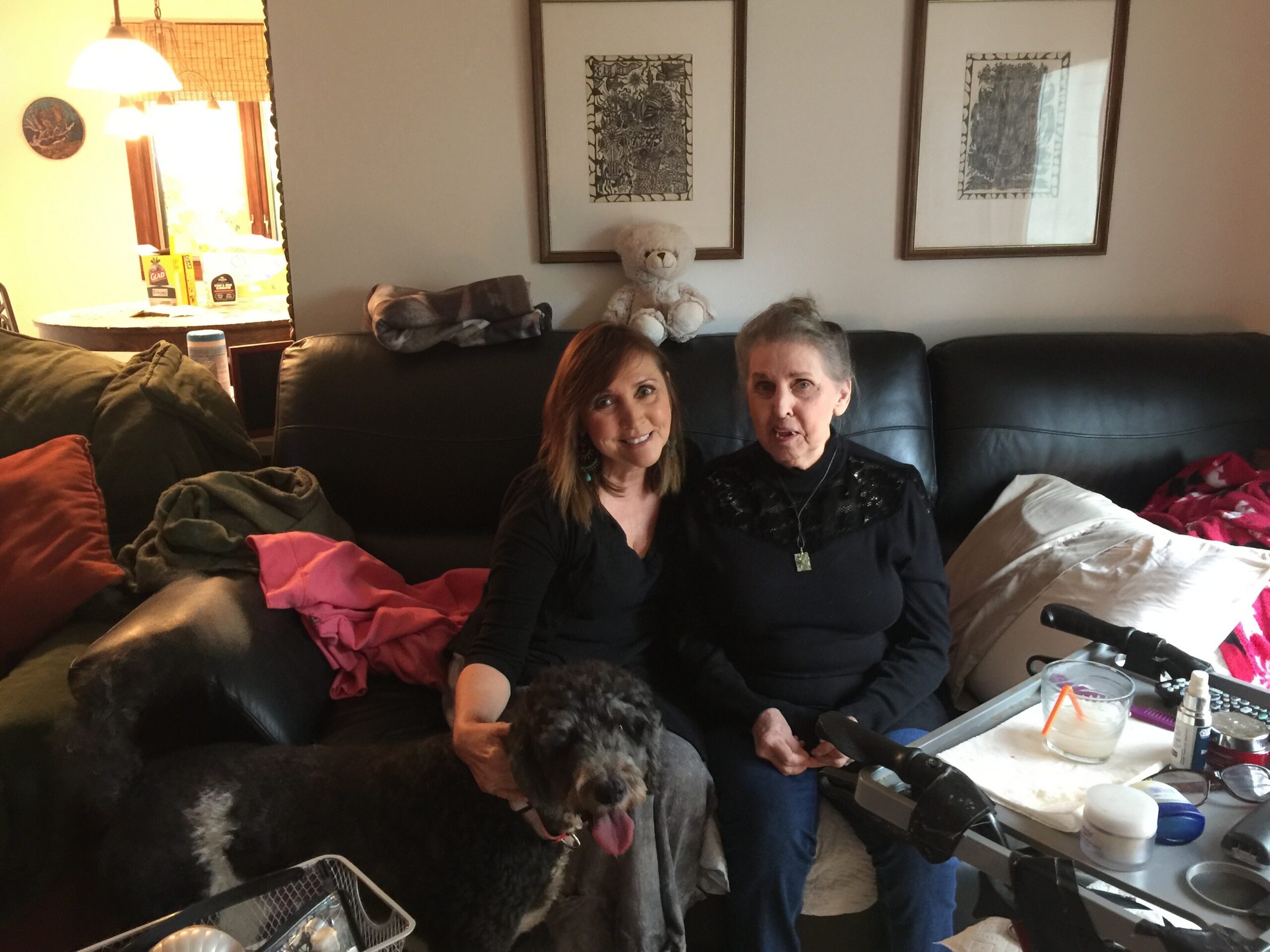 Two women sit on a black leather couch with a fluffy poodle in front, in a cozy living room with framed wall art behind them.