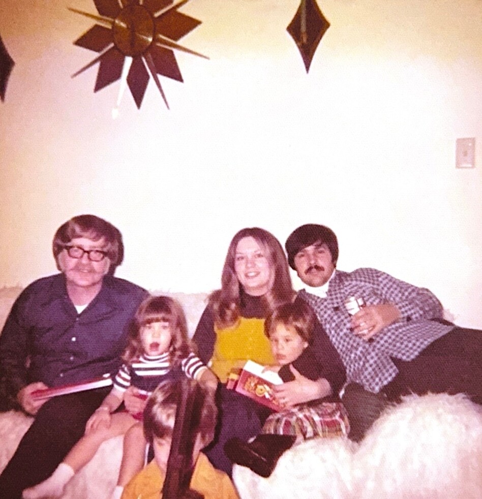Living room family photo: two adults on a couch with three children, one child sitting on the floor in front.