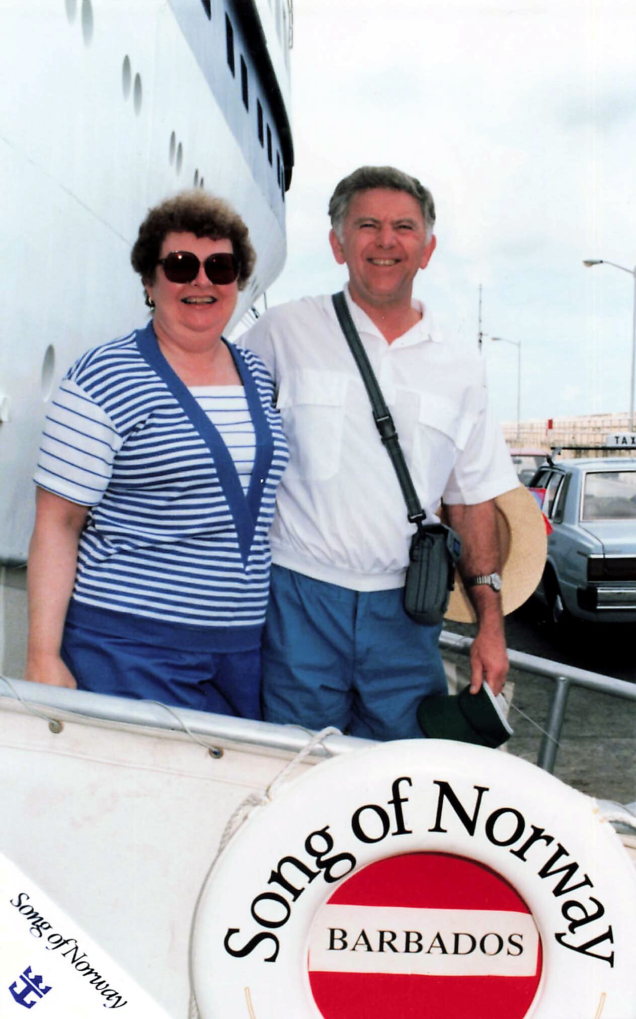 Two smiling tourists posing in front of a large white ship with a circular buoy reading 'Song of Norway Barbados' in the foreground.