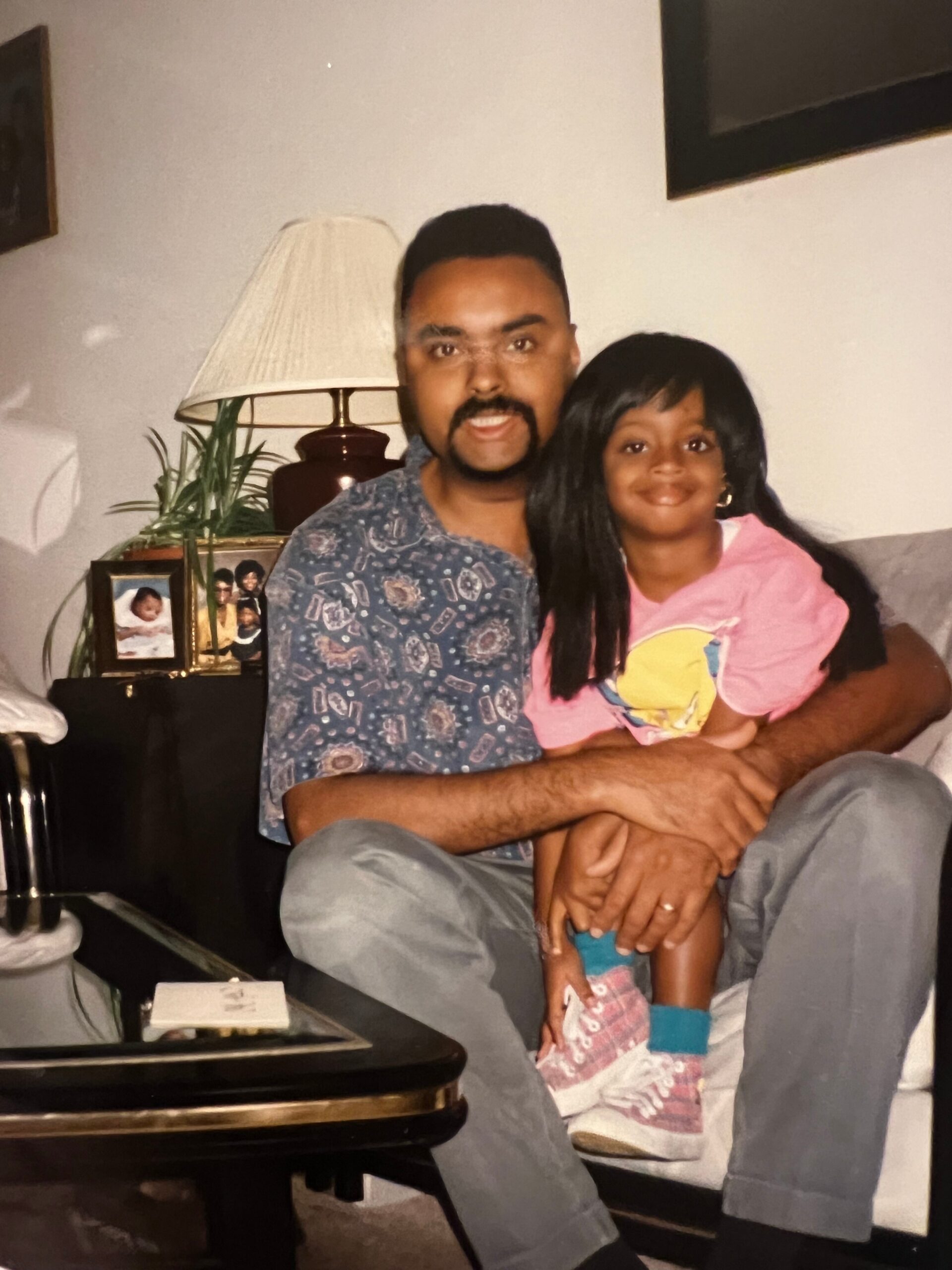 Man with a mustache sitting on a couch holding a smiling girl in a pink shirt; cozy living room background with a lamp and family photos.