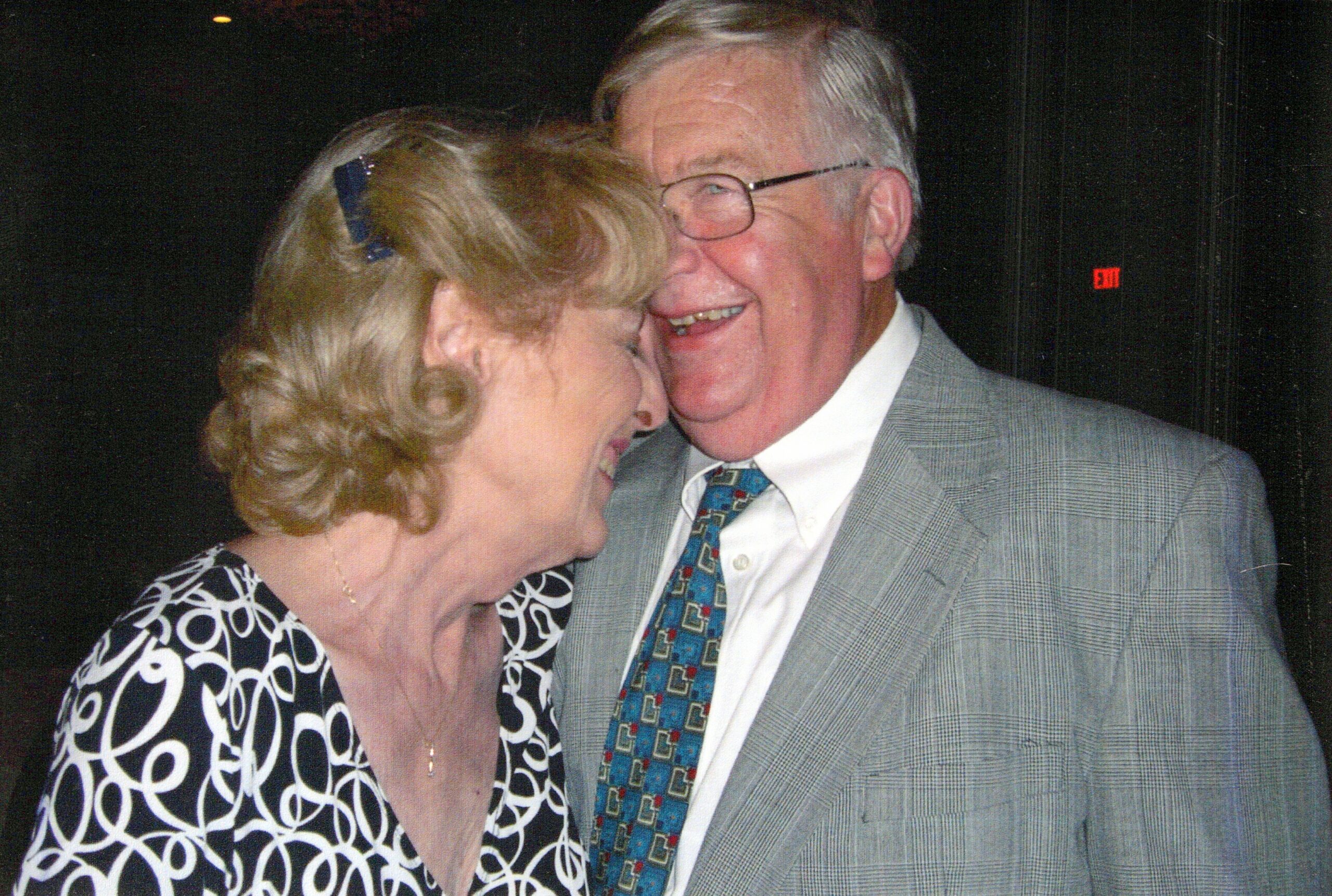 Elderly couple sharing a kiss on the cheek at a formal event, both smiling.