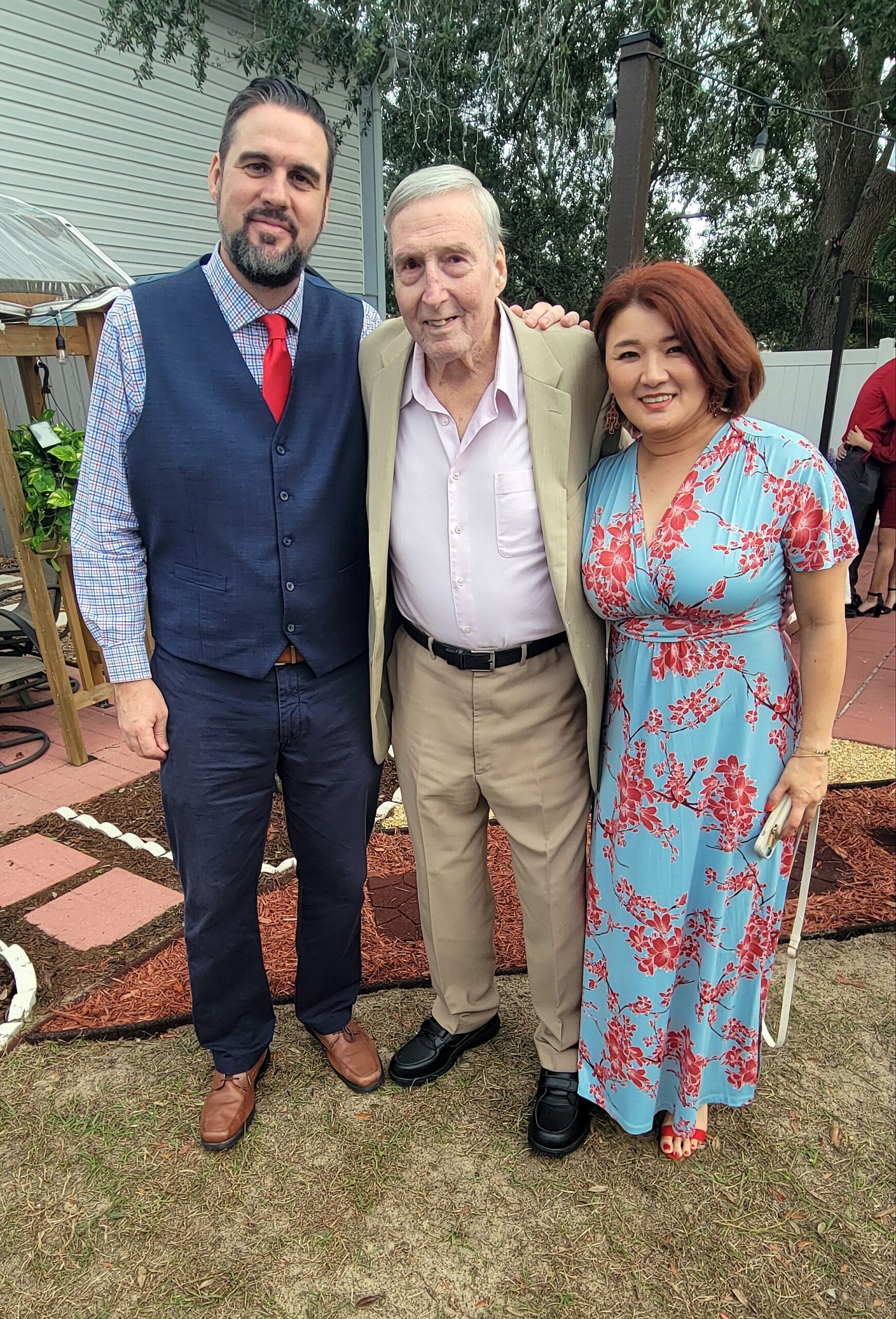Three adults posing together outdoors at a backyard party: man in a navy vest with red tie, an older man in a beige suit, and a woman in a blue floral dress smiling at the camera.