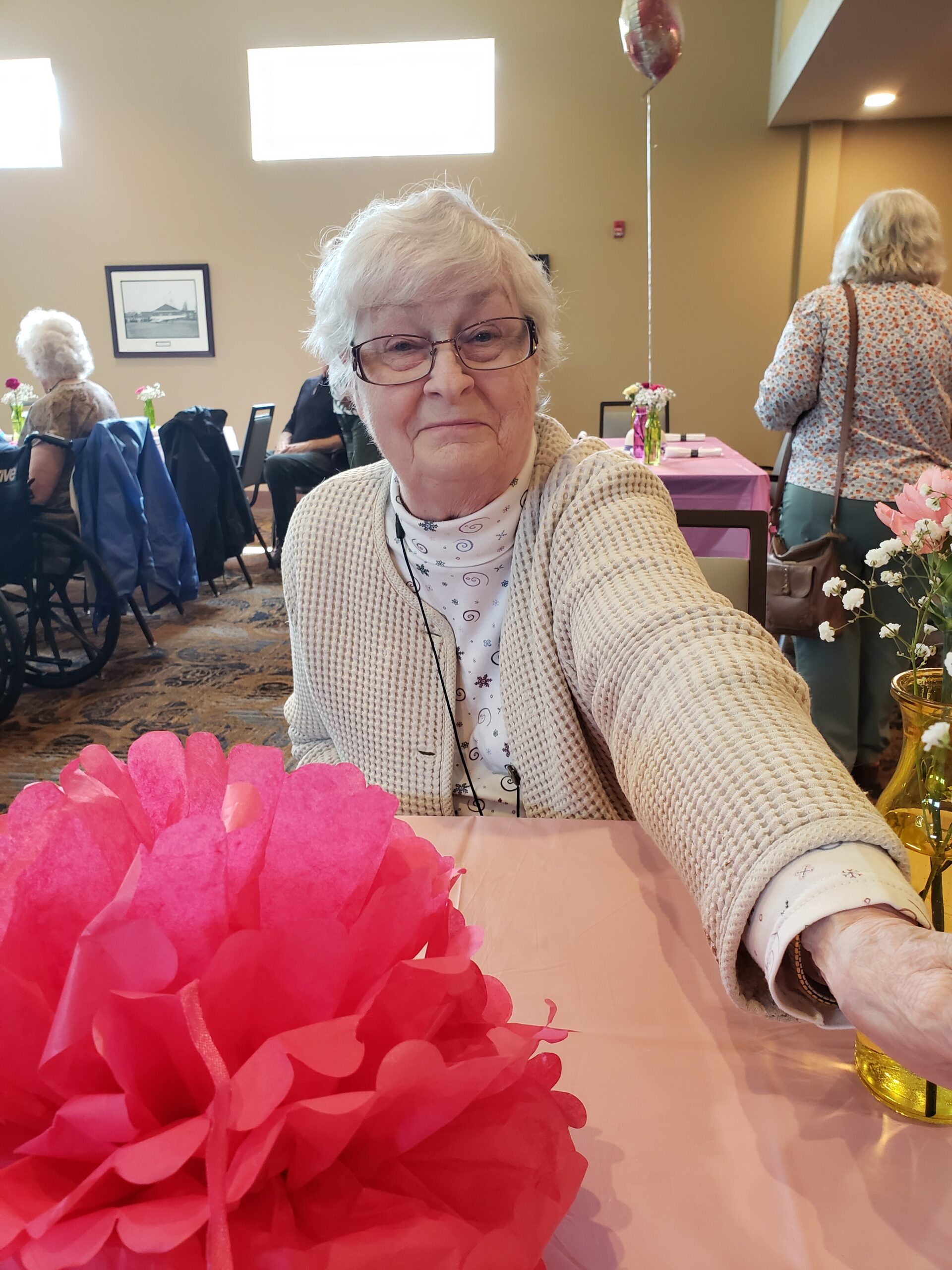 Senior woman with white hair and glasses takes a selfie at a celebration, pink tissue-paper centerpieces and pastel decor in the foreground.