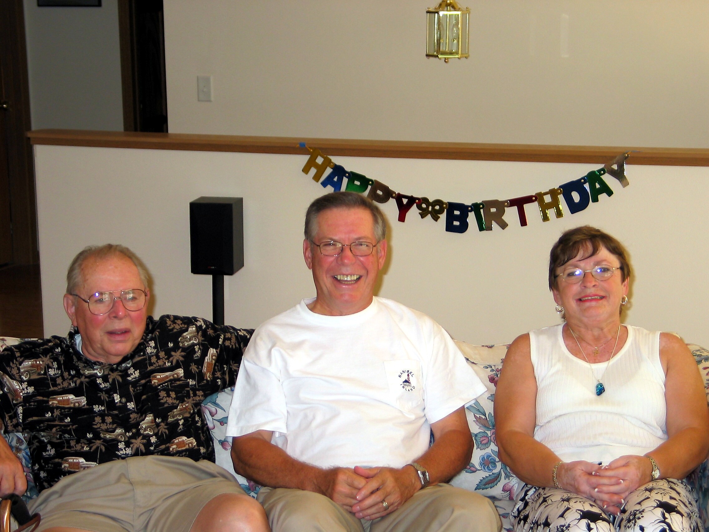 Three smiling seniors sit together on a couch at a birthday party, with a colorful 'Happy Birthday' banner in the background.