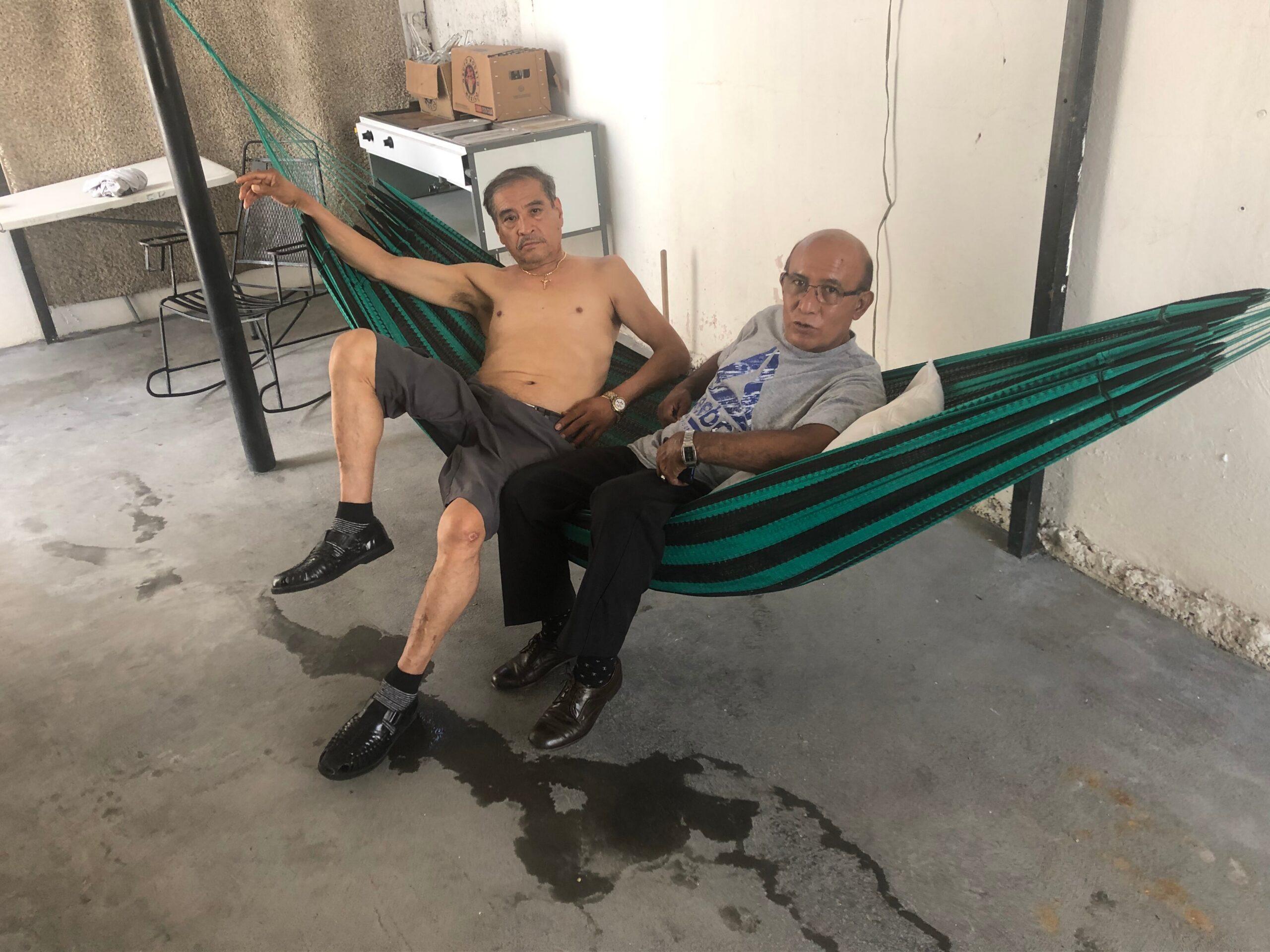 Two men lounging in a green striped hammock indoors; one shirtless, the other in a gray shirt and glasses, with a spill on the concrete floor nearby.
