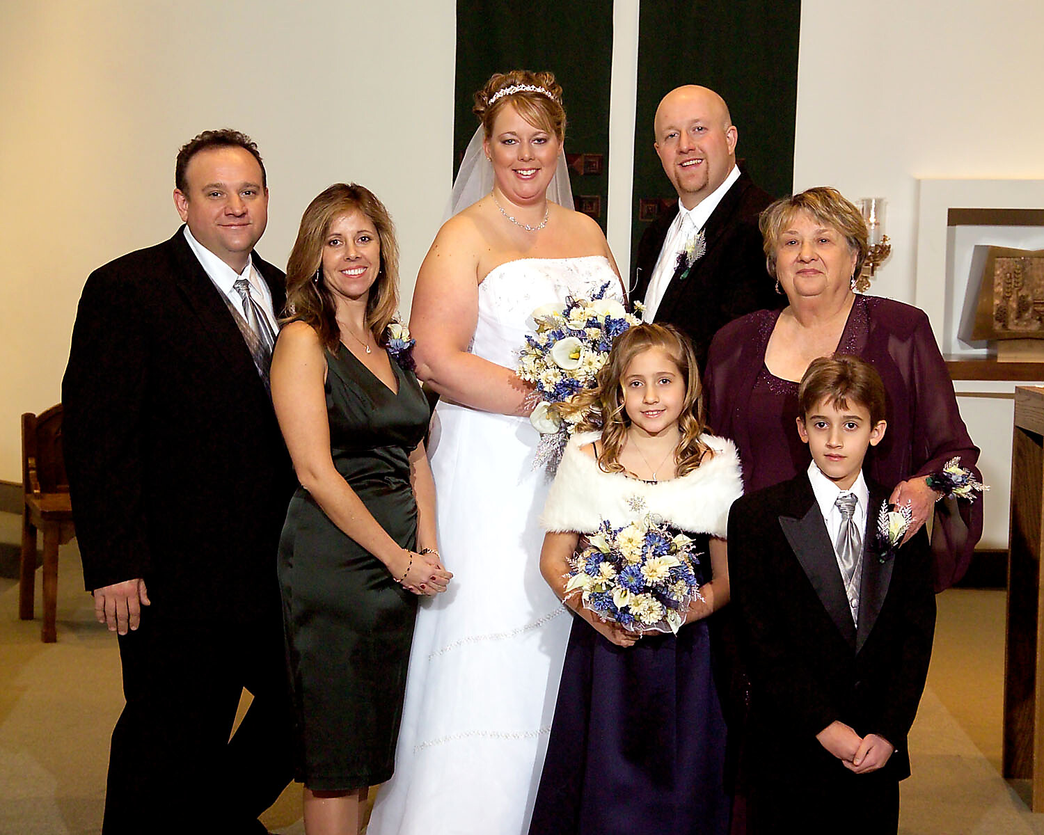 Bride in a white gown and veil, holding a bouquet, flanked by family at a wedding photo.