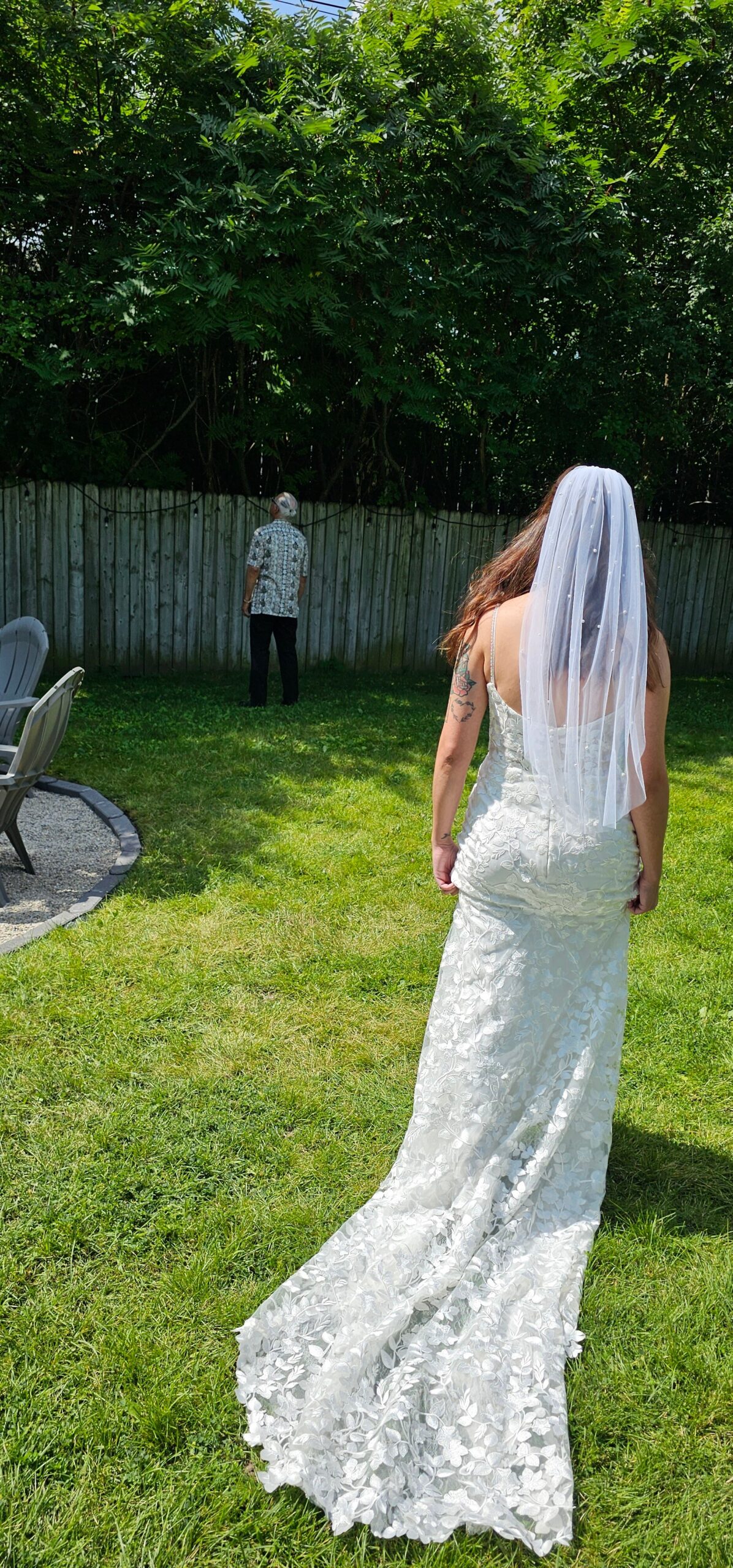Bride in a white lace wedding dress with a veil, seen from behind, walking toward a man near a wooden fence in a sunny backyard garden.