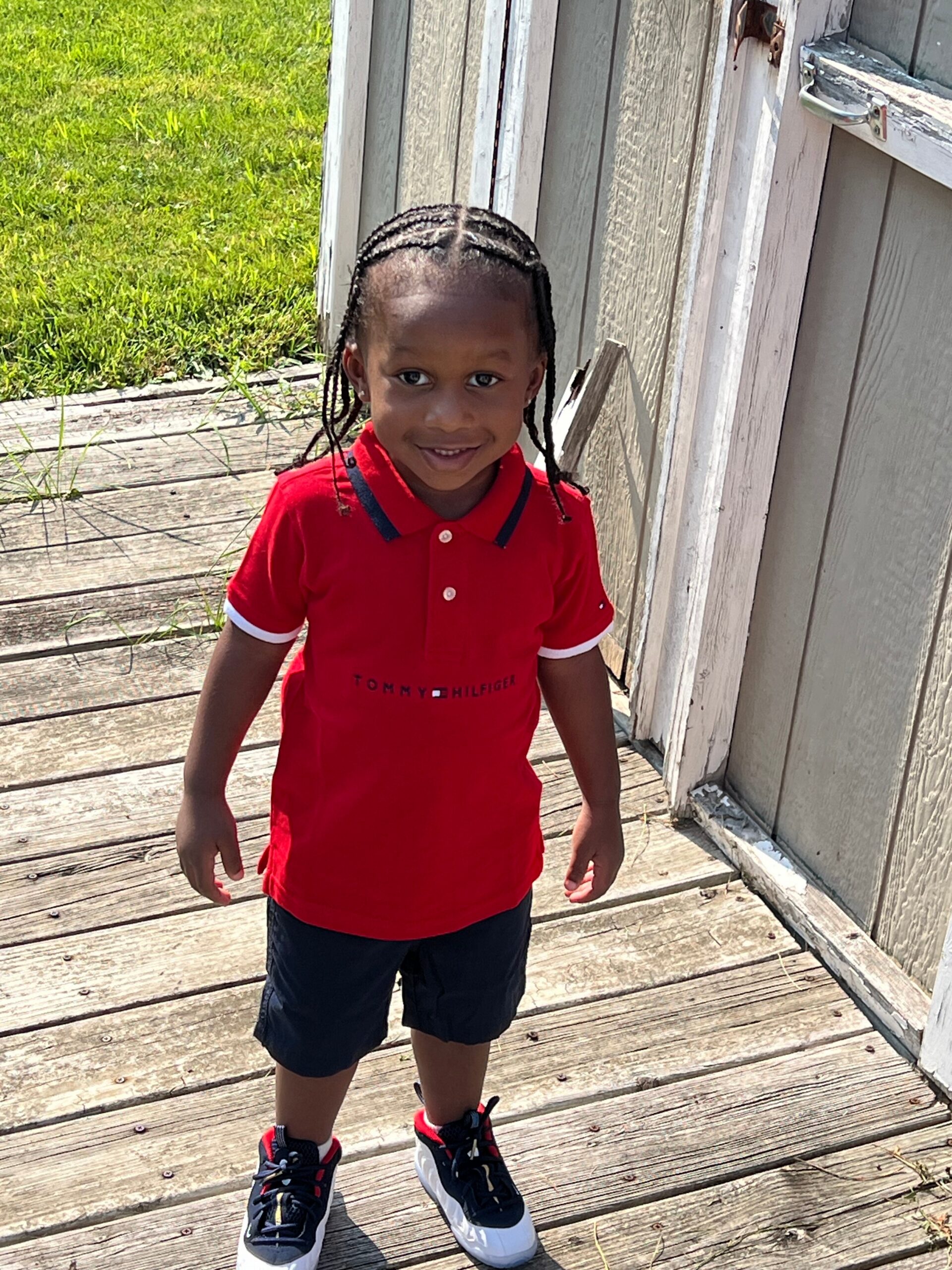 Young child standing on a wooden deck outside, wearing a red polo shirt and black shorts, sneakers, smiling at the camera.