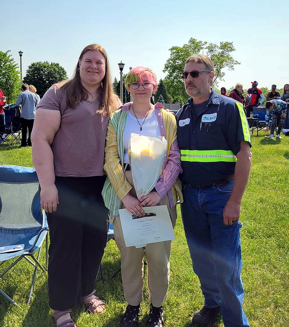 Outdoor award moment: a young person with pink hair holds a bouquet and certificate, flanked by two adults at a sunny grassy event.
