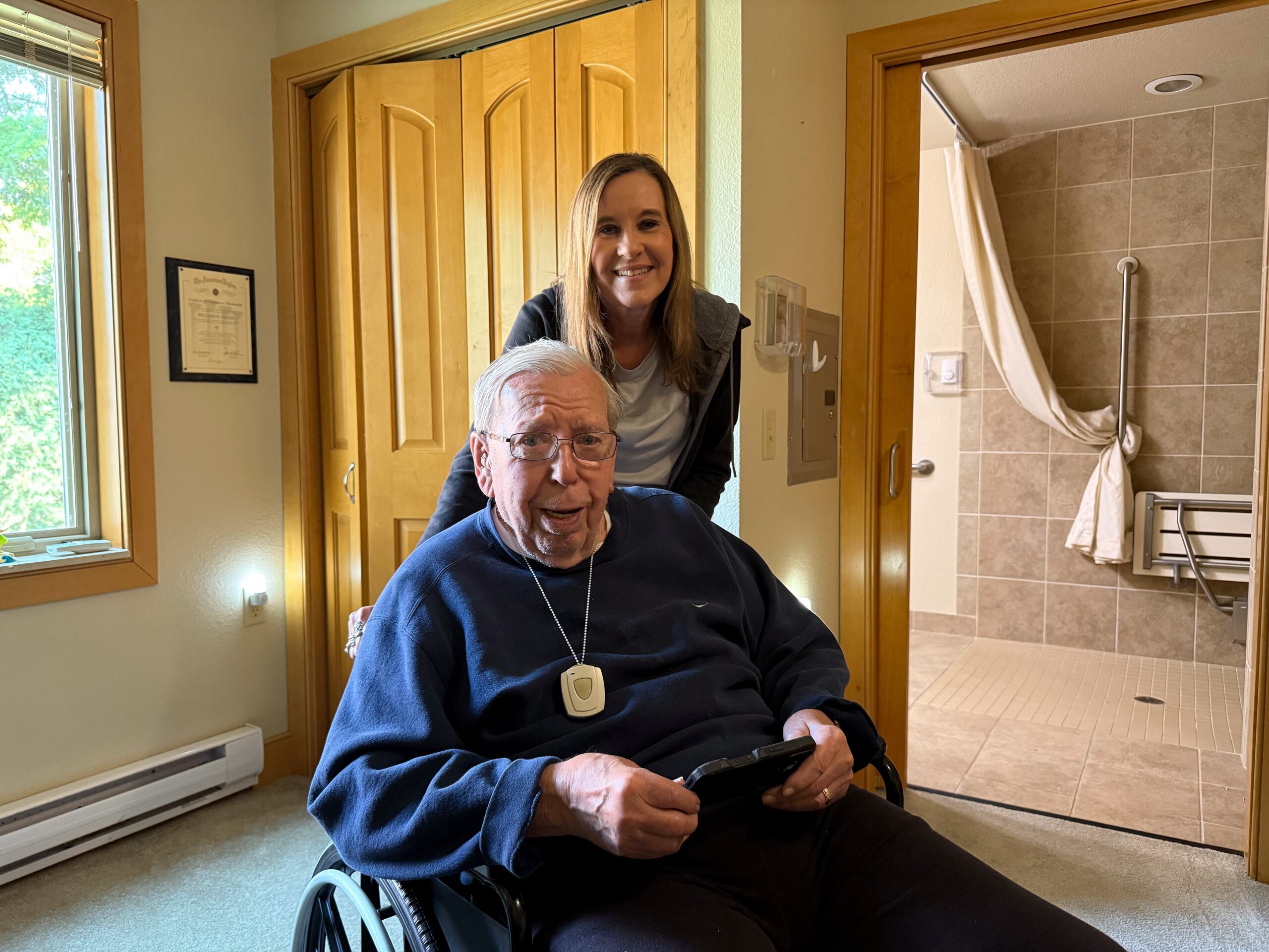 An elderly man in a wheelchair smiling, with a caregiver standing behind him in a doorway of a warm, homey hallway.