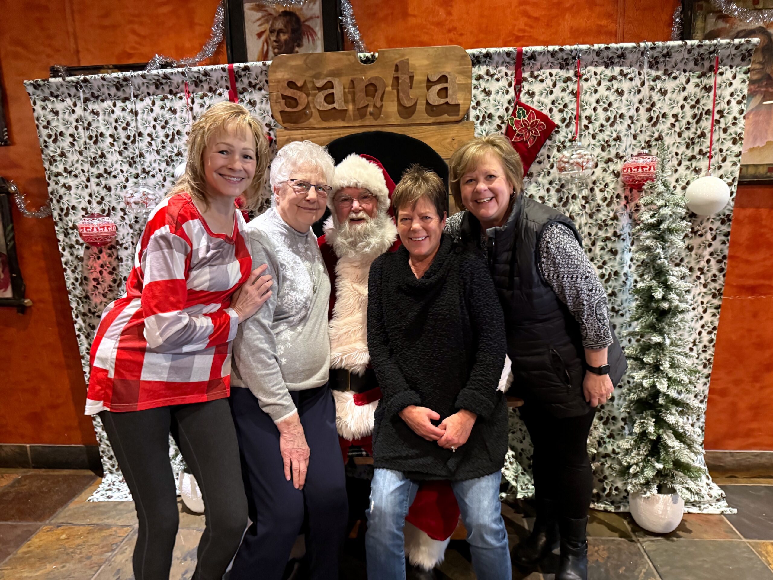 Group of five adults posing with Santa Claus in a festive holiday setting and patterned backdrop.