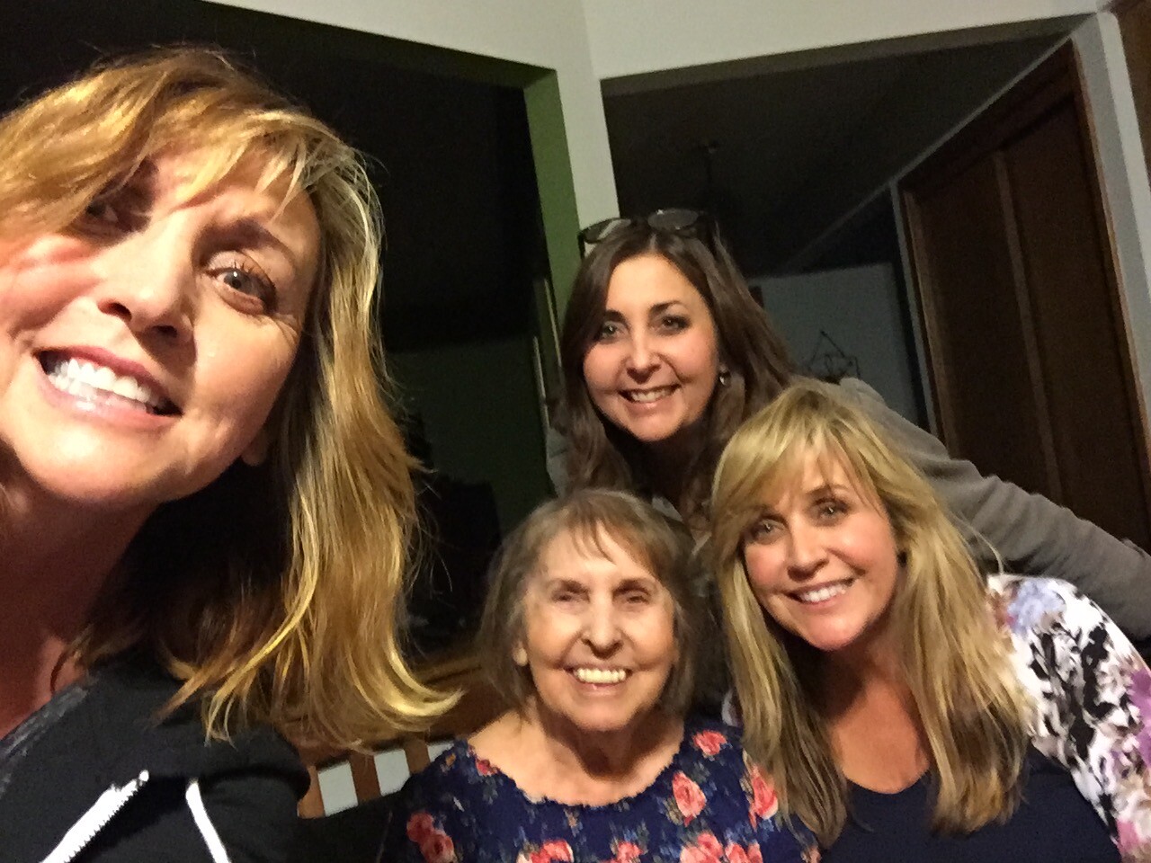 Four women are smiling for a group selfie indoors, with one in the foreground on the left and three others behind them.
