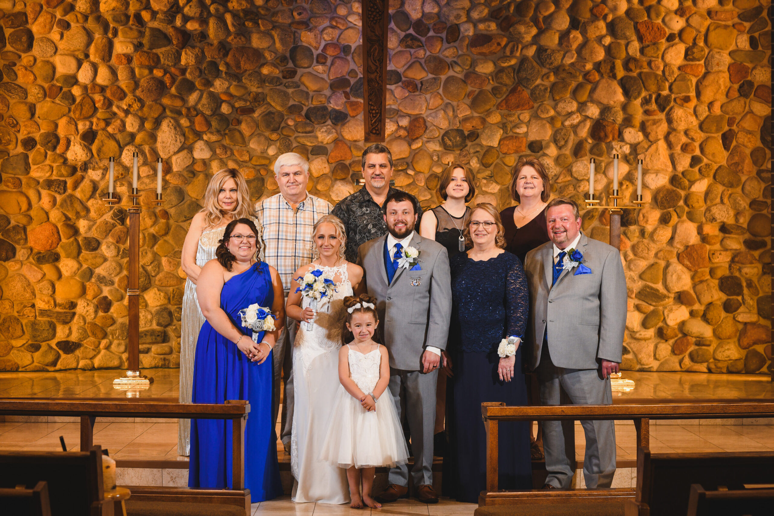 Group of ten adults and two children posing for a wedding party photo in a stone chapel backdrop. Center: bride in white, groom in gray suit, others in formal attire.