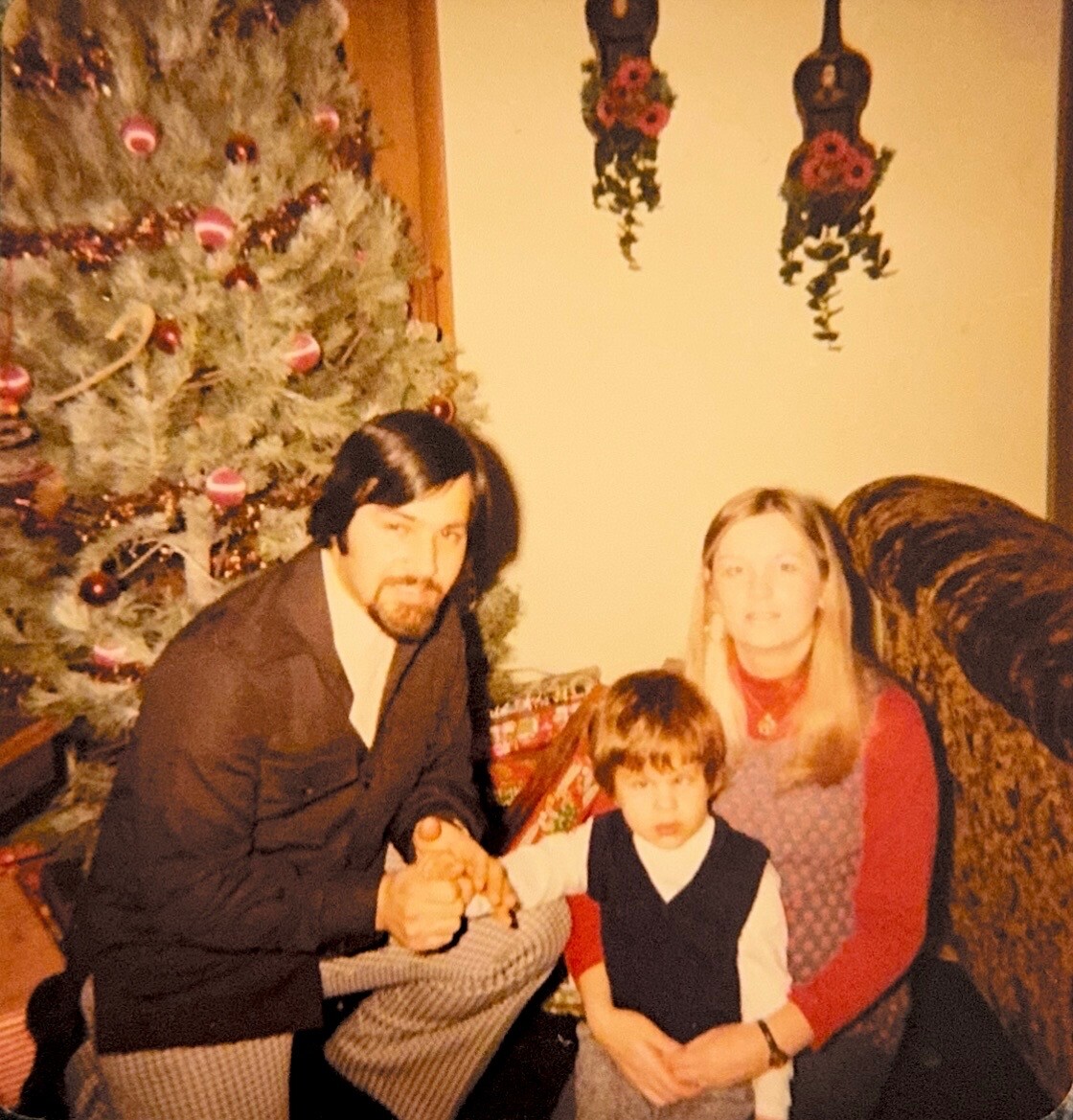 A man with dark hair and a beard kneels beside a young boy and a woman on a living room floor, with a decorated Christmas tree in the background.