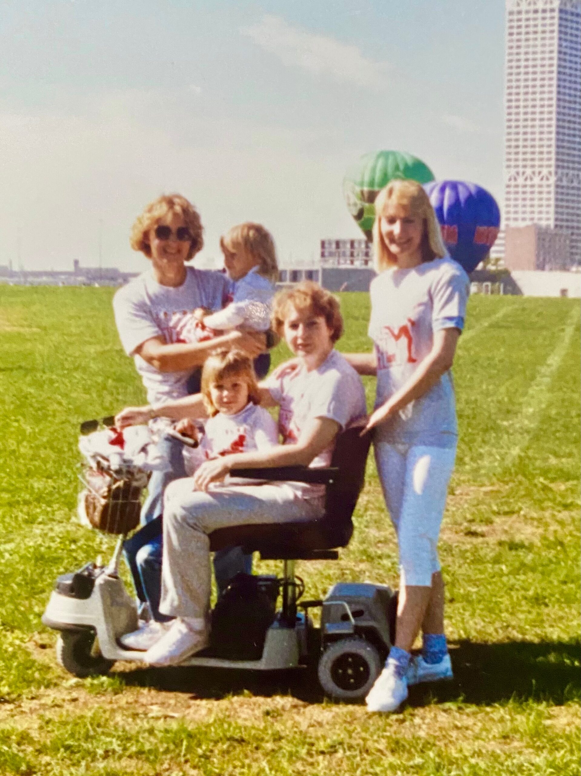 Family posing in a grassy field on a sunny day; a person in a motorized wheelchair sits front and center with others standing nearby, colorful balloons in the background.
