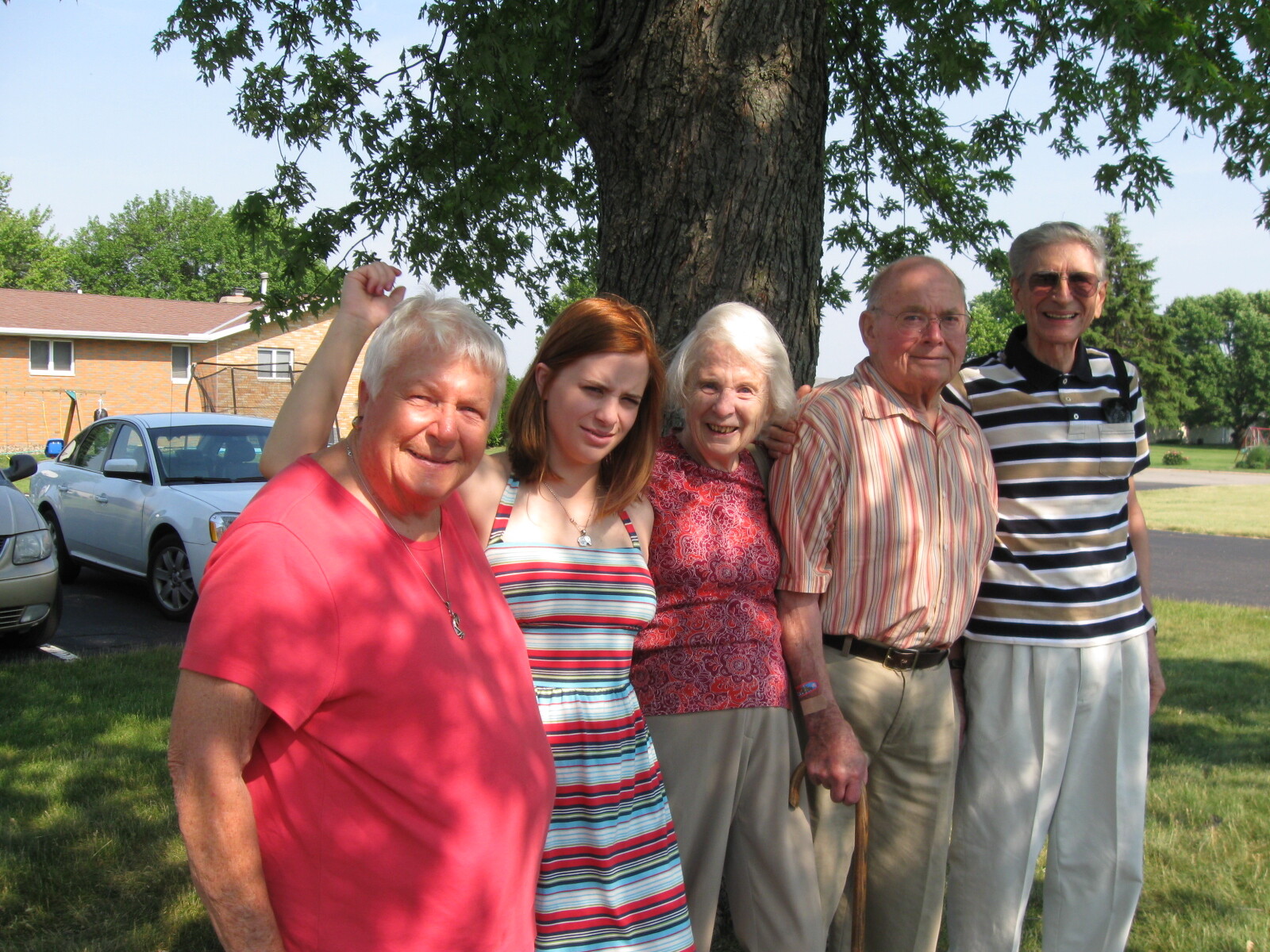 Five generations/family members posing together under a large tree outdoors, smiling at the camera.