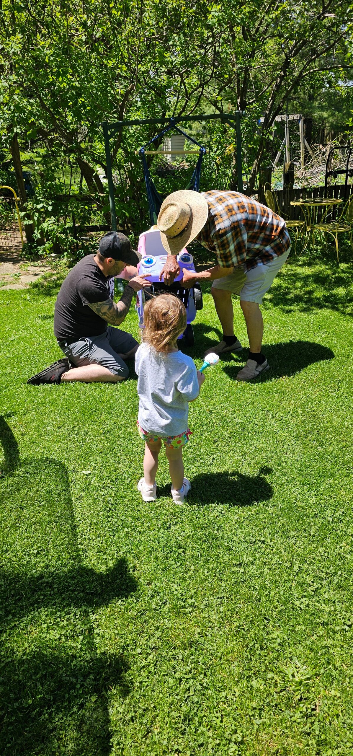 Two adults kneel in a sunny backyard, fixing a blue and purple ride-on toy for a young child as another child watches with a bubble wand in hand.
