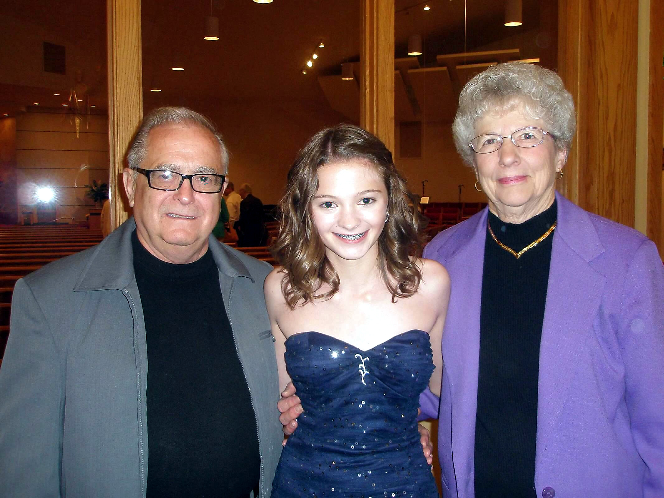 Three generations posing together indoors: an elderly man on the left, a teenage girl in a blue dress in the center, and an elderly woman in a purple jacket on the right.
