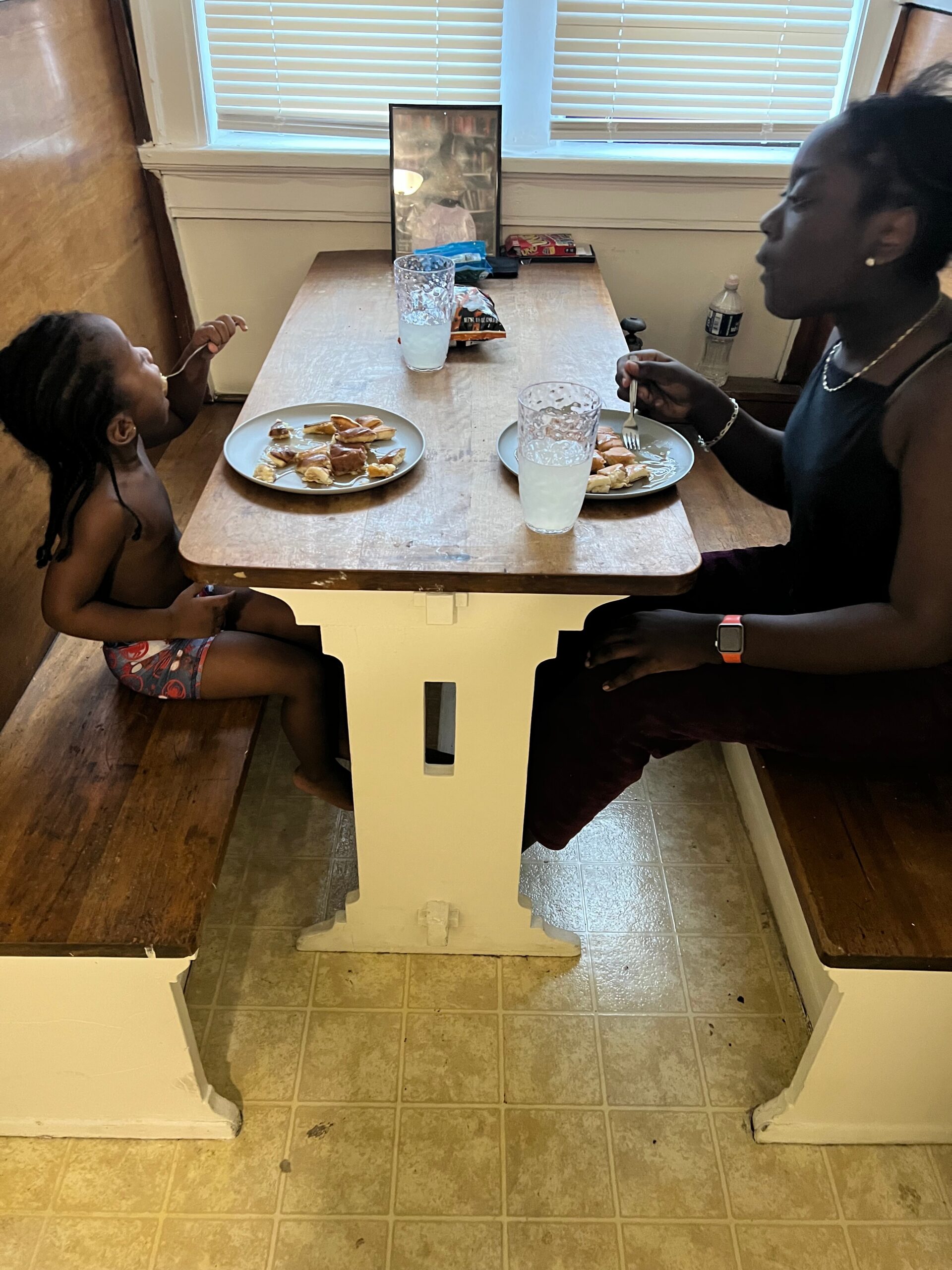 Mother and child sit at a small white table eating pastries on white plates, with milk in tall glasses, in a kitchen nook by a window.