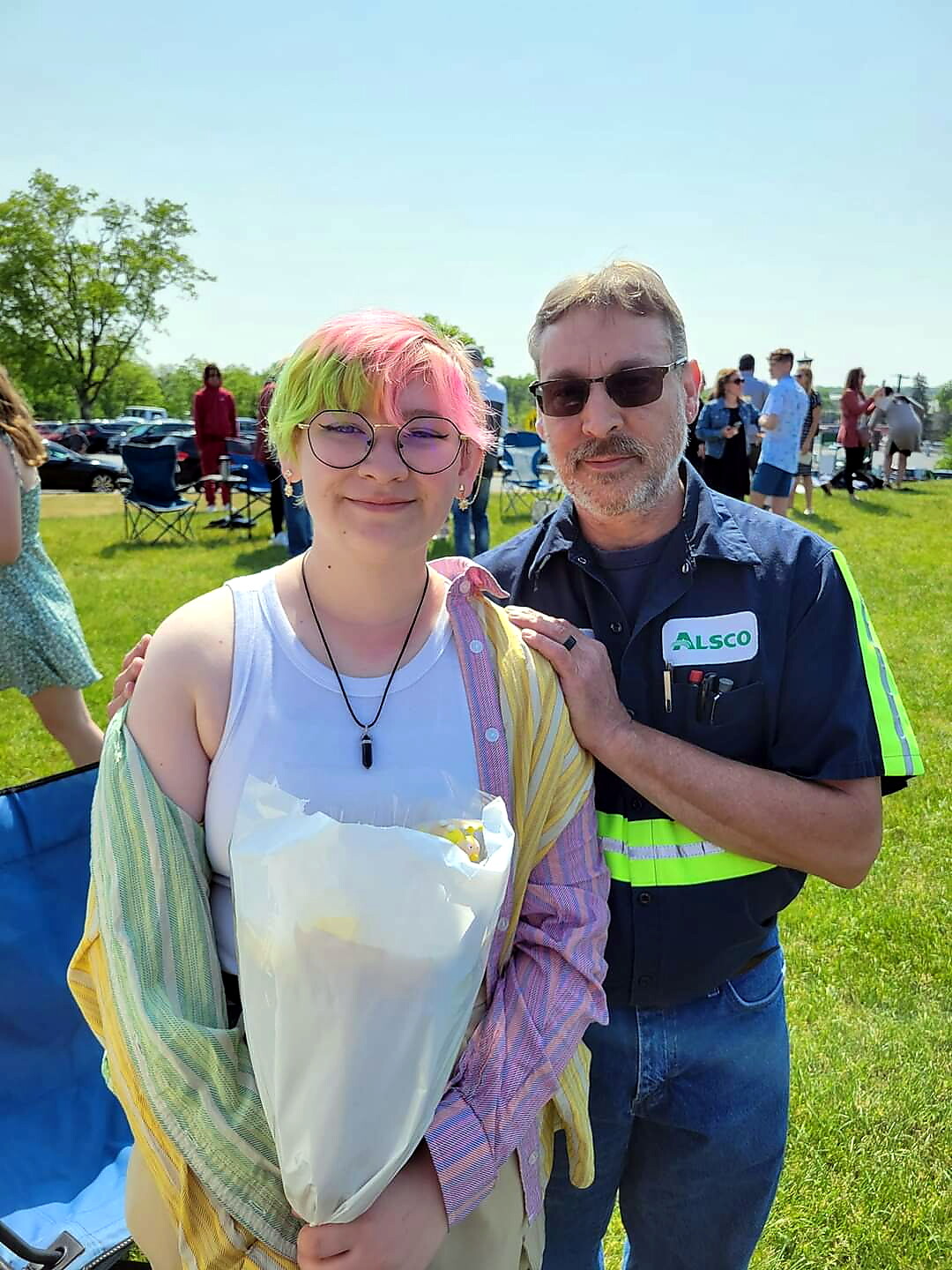 Young person with pink‑green hair and glasses holding a wrapped item, posing with a man in a blue ALSCO uniform at an outdoor event on a sunny day.
