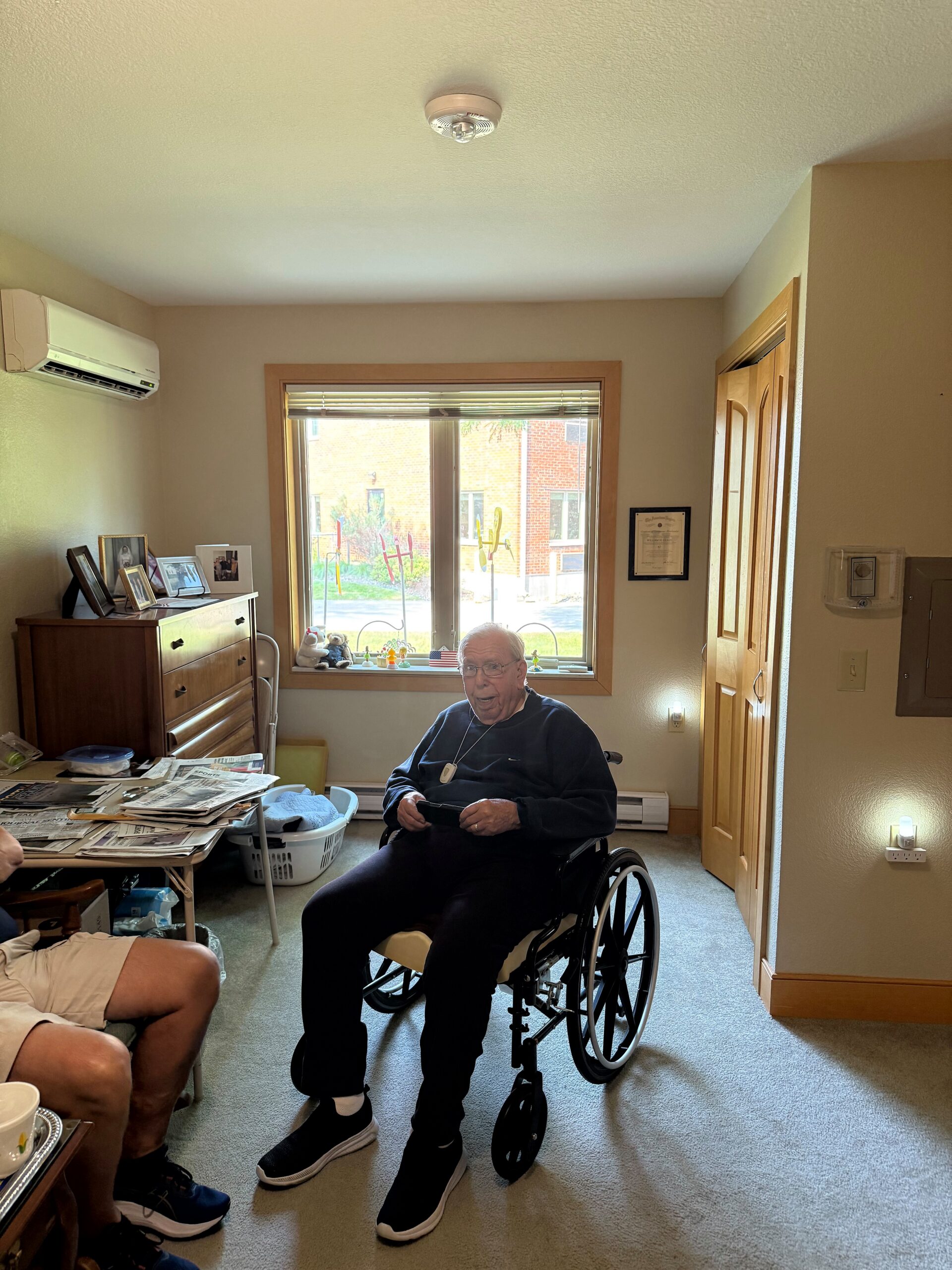 Elderly man in a wheelchair sits in a sunlit living room with a window and cluttered table to the left.