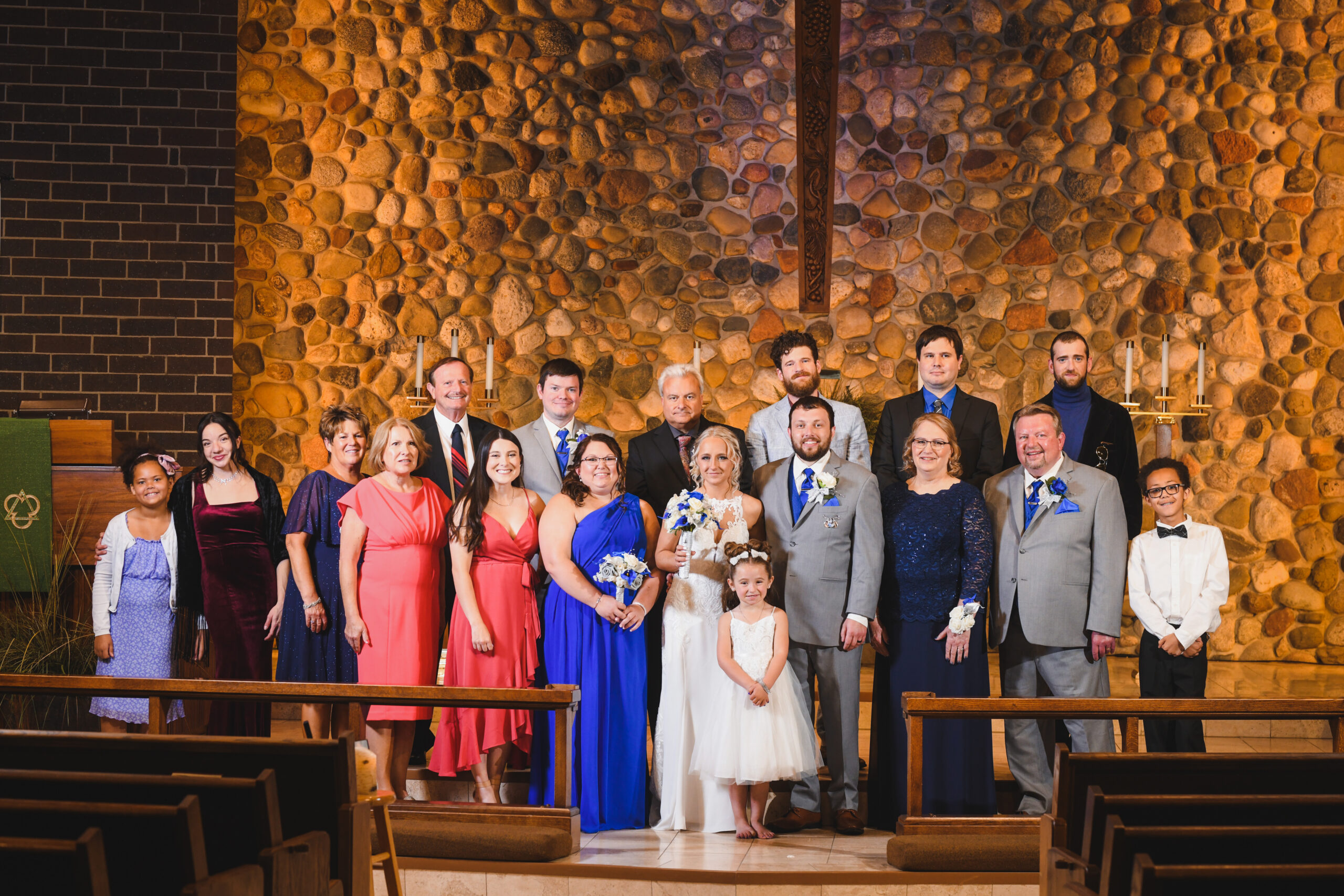 Group of wedding guests and two brides in gowns posing on the church steps in front of a stone wall backdrop.