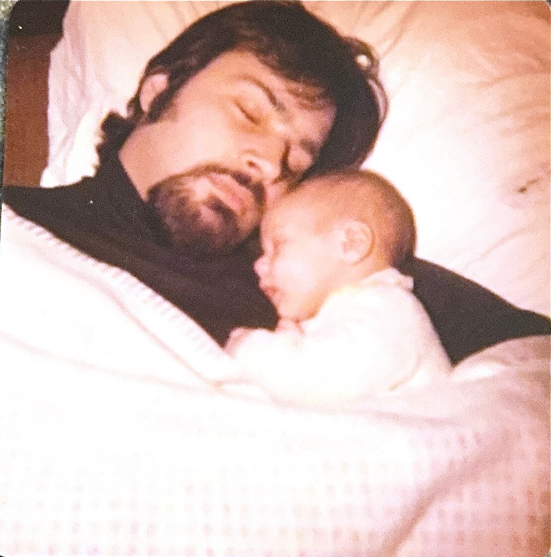 Man with a beard asleep in bed, cuddling a newborn baby on his chest.