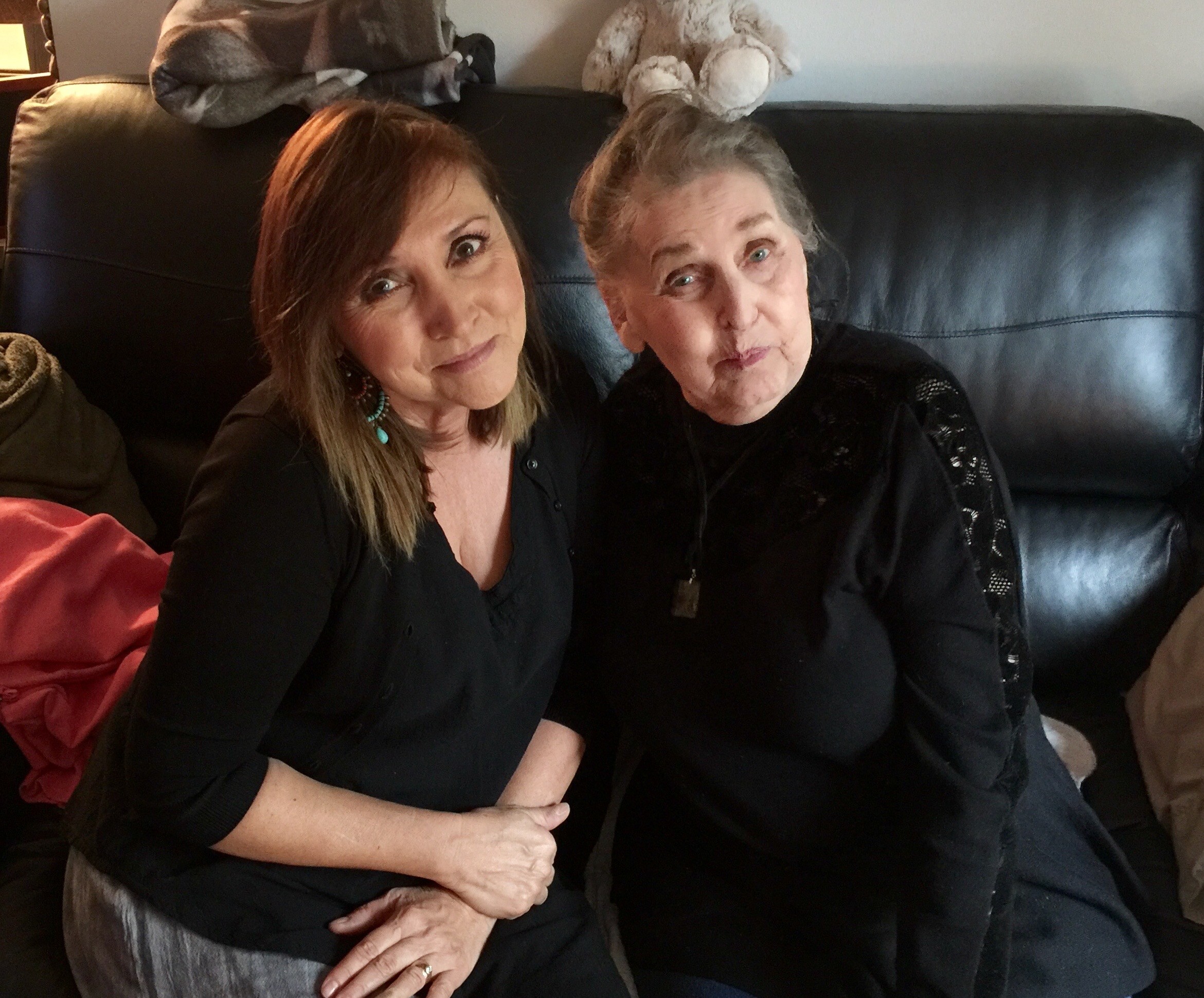 Two women sit together on a black leather sofa, smiling for a selfie on a couch.