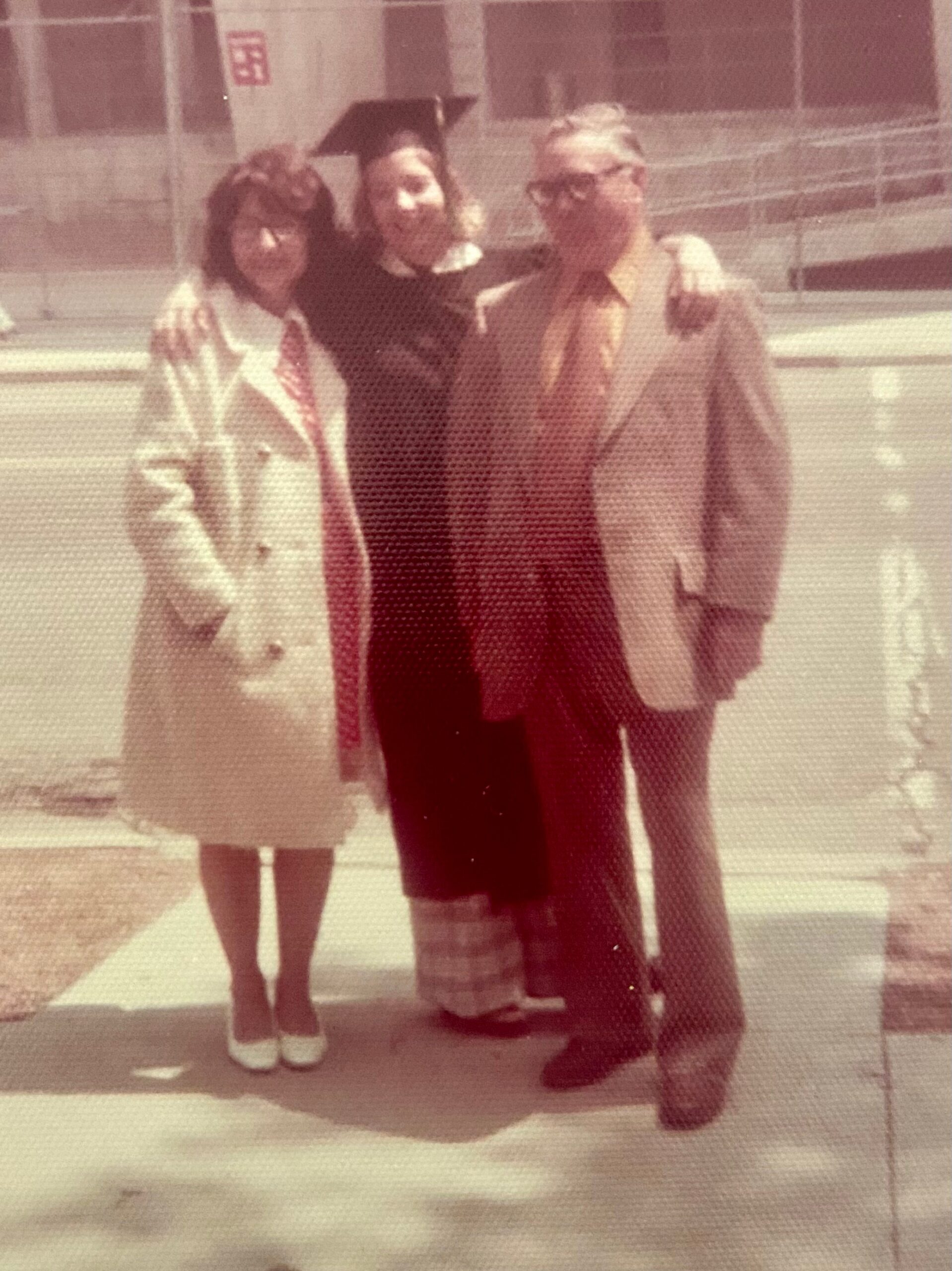 Three people stand together for a graduation photo: a graduate in a cap and gown between two friends, one in a light coat and the other in a tan jacket.