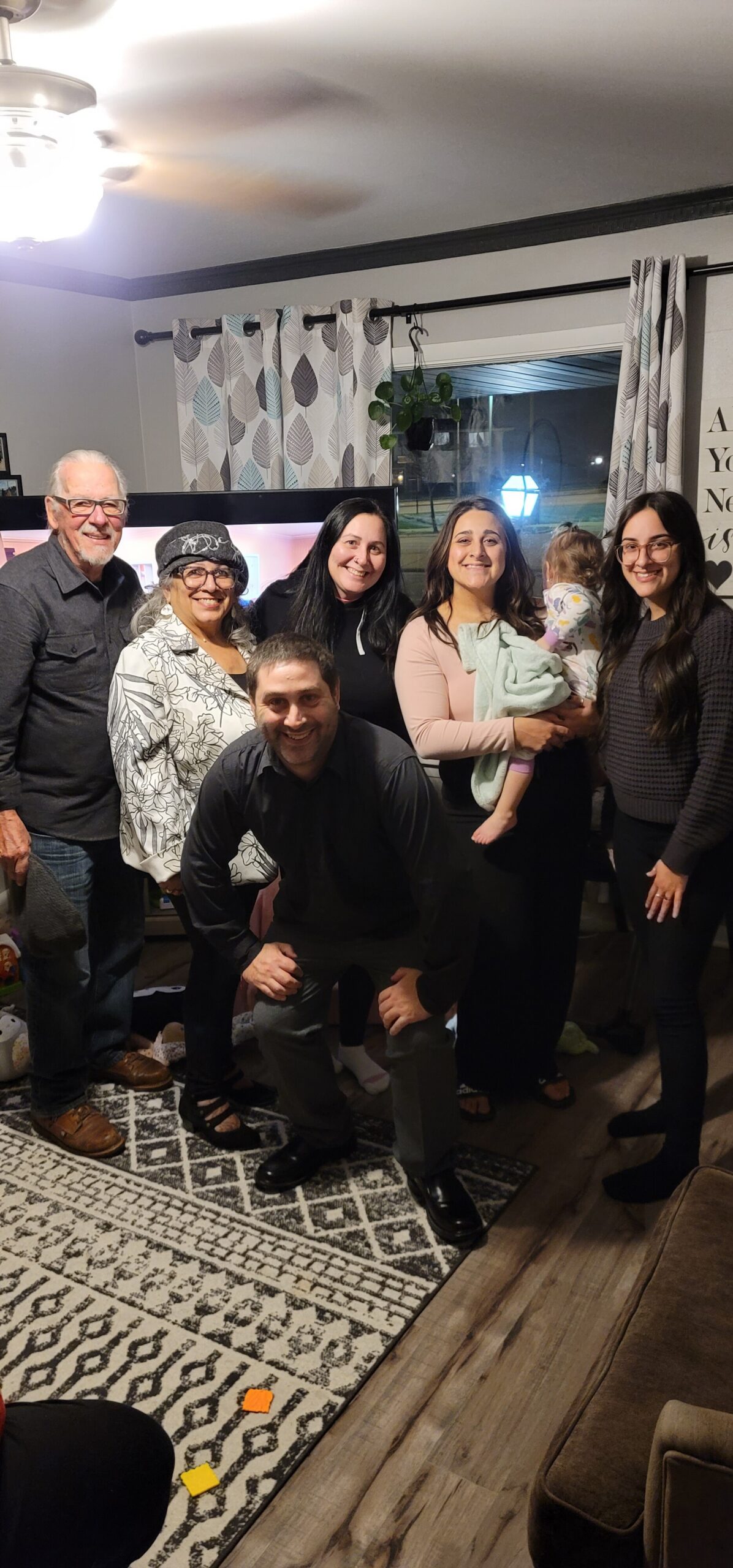 A group of six adults standing in a living room with a baby, smiling for a casual family photo near a patterned rug and curtains.