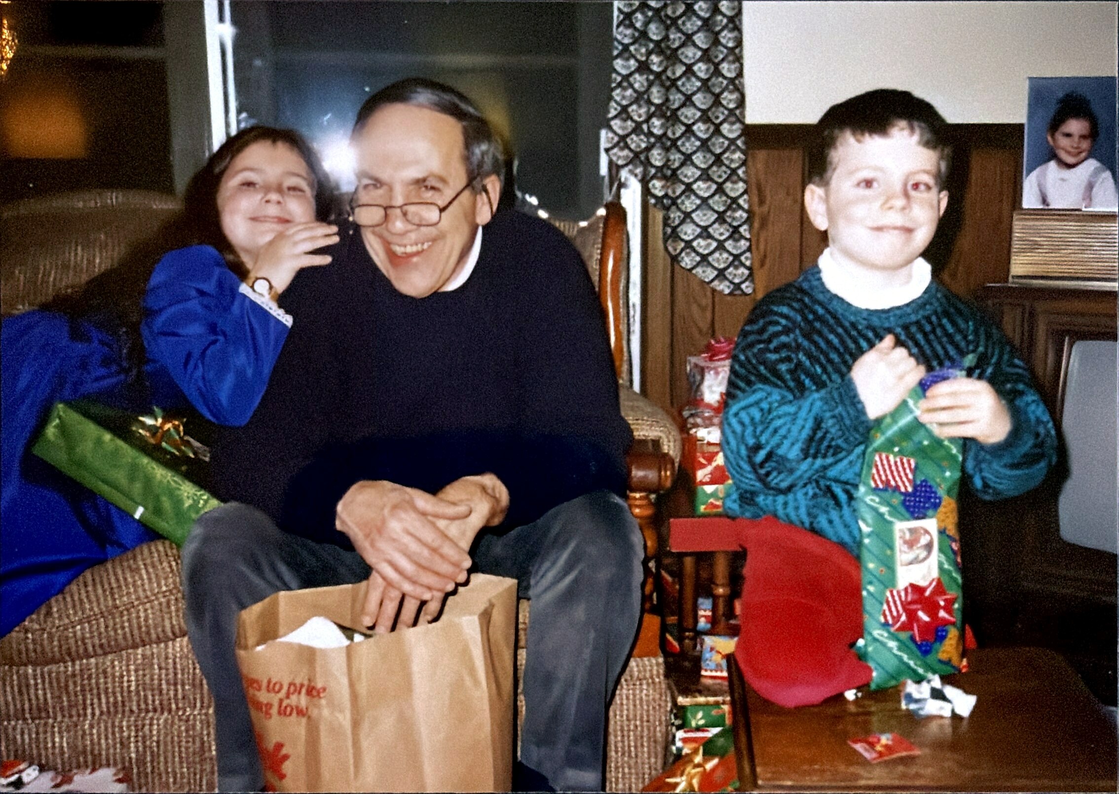 A smiling elderly man with reading glasses sitting between a girl and a boy during a gift exchange at home, wrapped gifts on the floor.