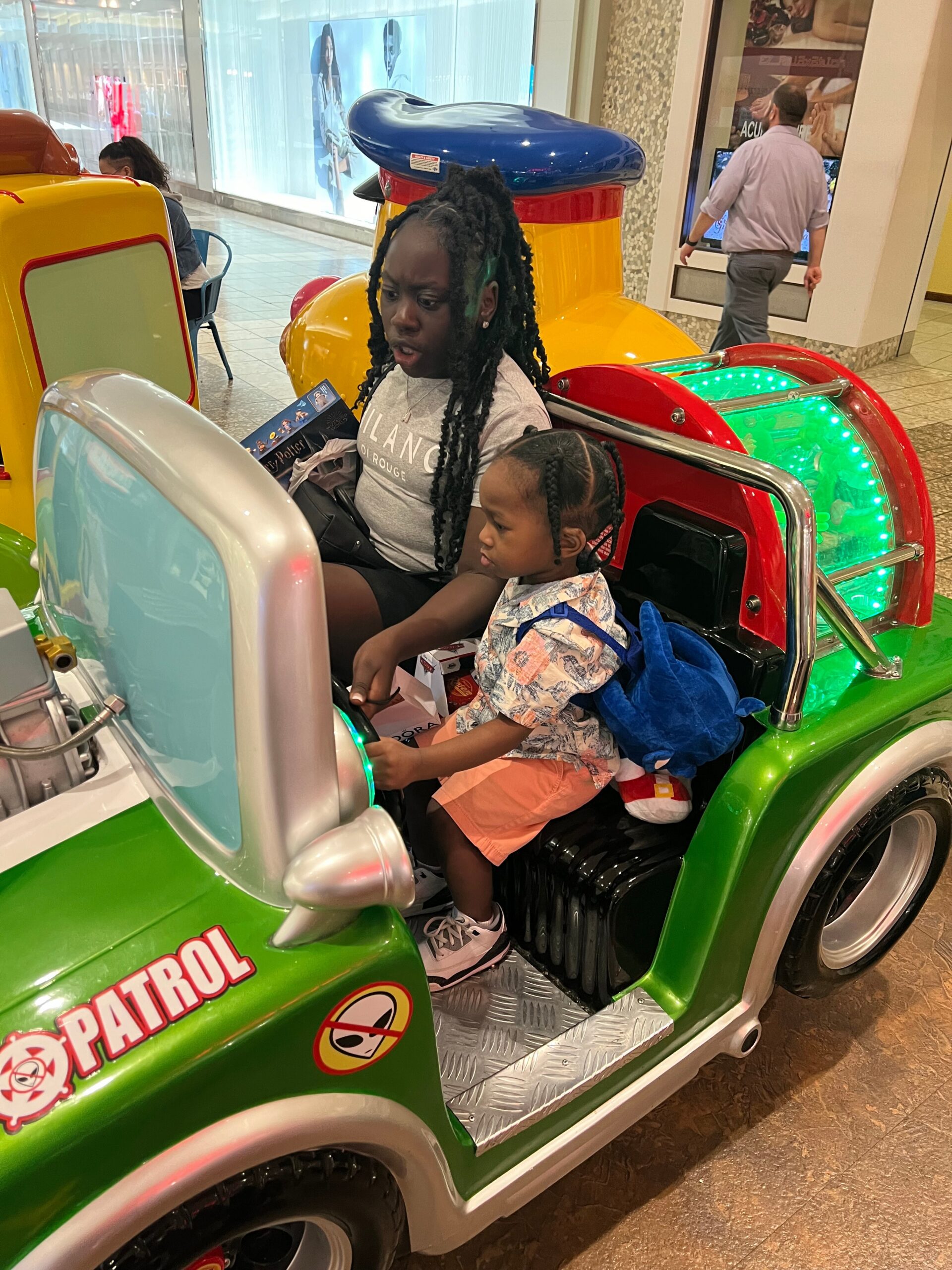 Two kids ride in a bright green toy police car in a mall, one steering while the other sits beside with a stuffed toy nearby.