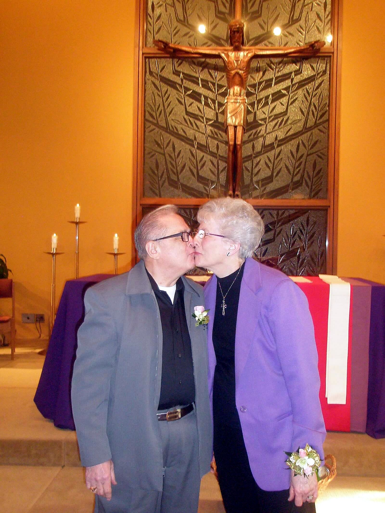 Elderly couple kissing in a church with a large crucifix and stained glass behind them.